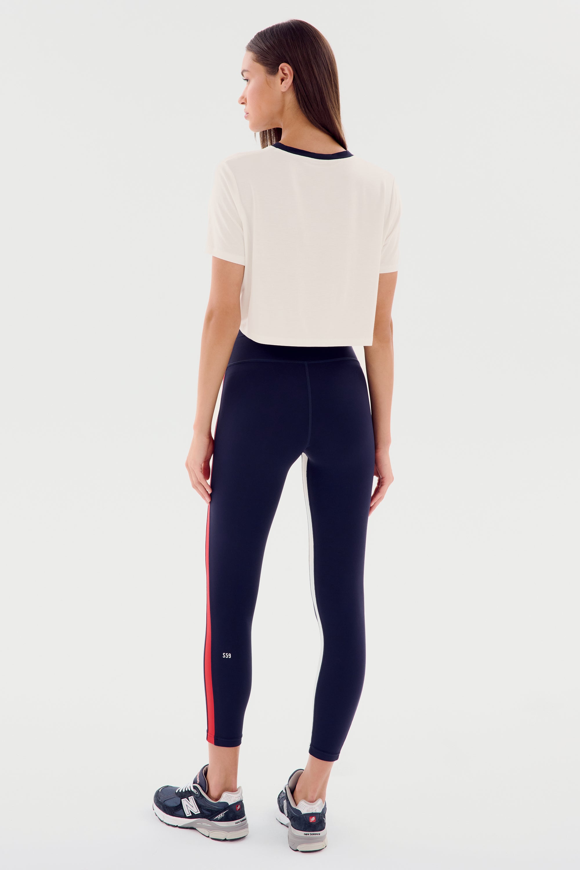 A person wearing a relaxed fit Djuna Crop Ringer - White/Indigo by SPLITS59 and navy athletic leggings with a single red stripe stands facing away, showing the back view of the outfit. Ideal for warm weather workouts, they also sport black and white athletic shoes.