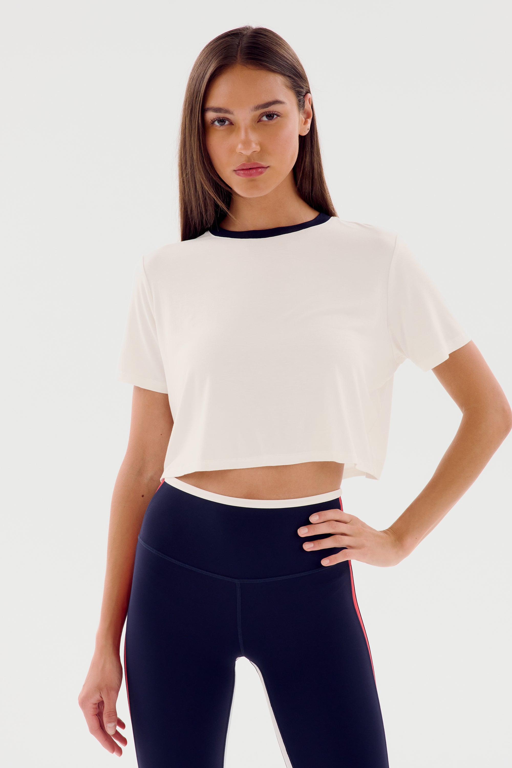 A woman with long straight hair wearing a white Djuna Crop Ringer by SPLITS59 and black leggings stands against a plain white background with her hand on her hip, ready for warm weather workouts.