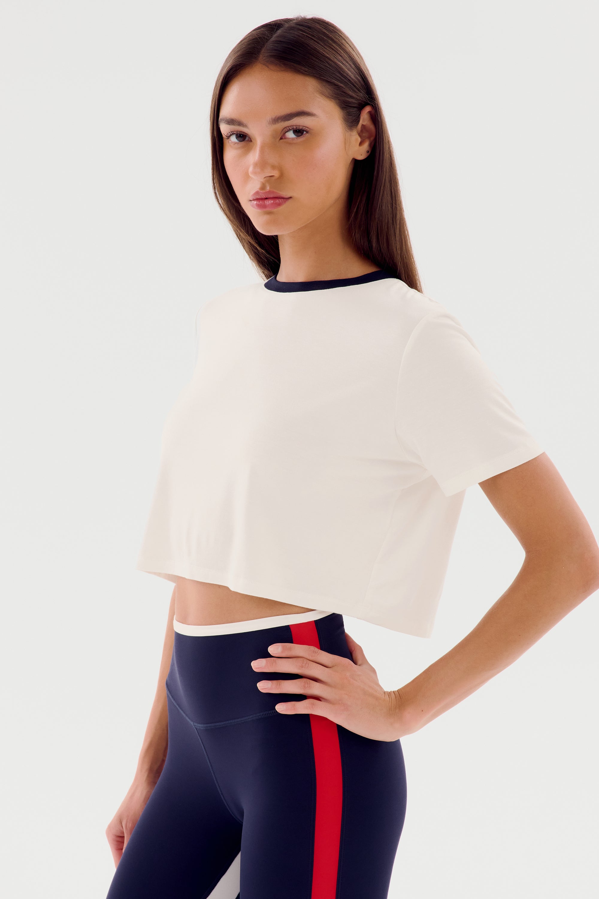 A person stands with one hand on hip, wearing a white relaxed fit **SPLITS59 Djuna Crop Ringer - White/Indigo** and navy leggings with red and white side stripes, perfect for gym workouts.