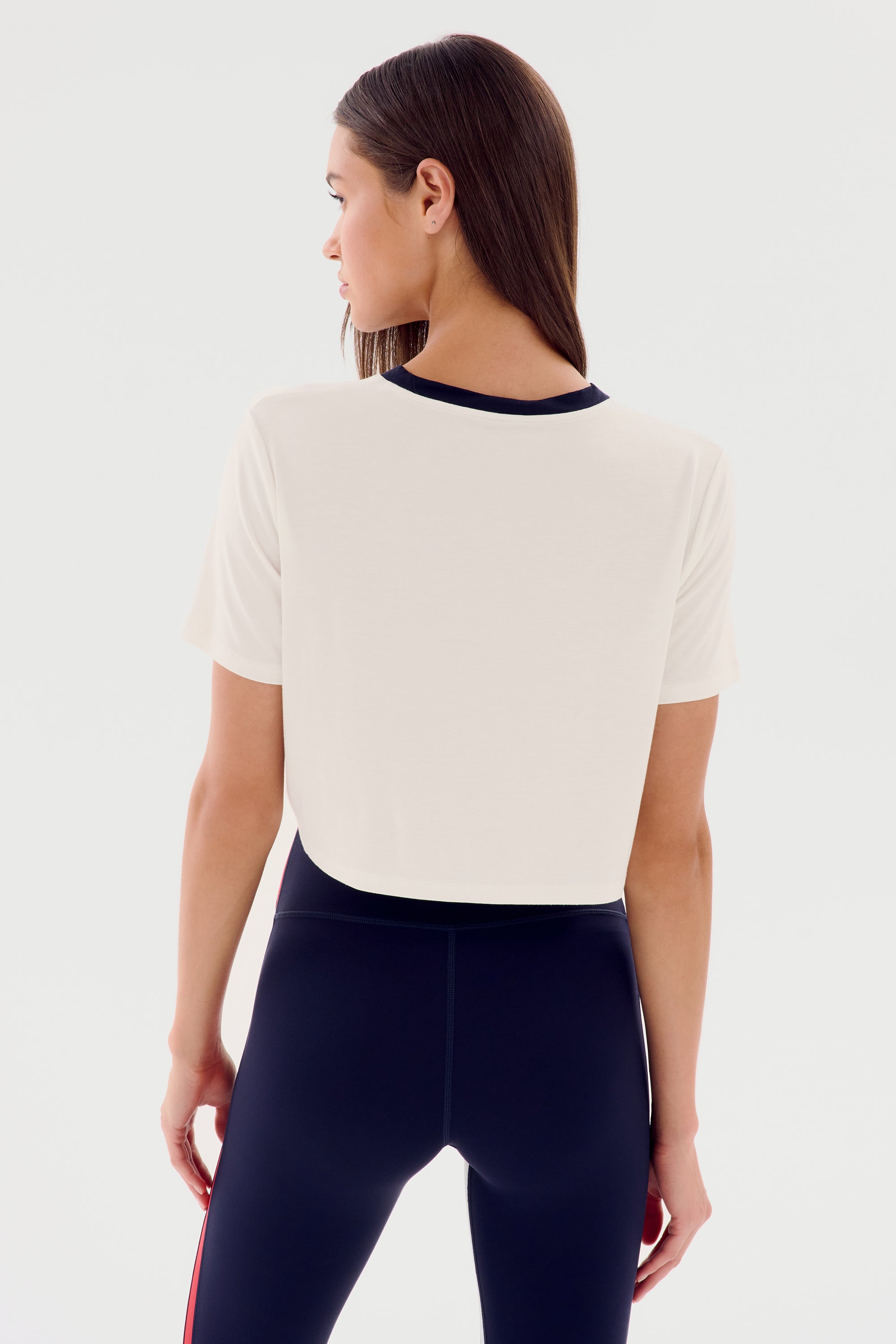 A person with long hair, wearing a SPLITS59 Djuna Crop Ringer - White/Indigo and black leggings with red stripes, is looking to the side, facing away from the camera against a plain background.