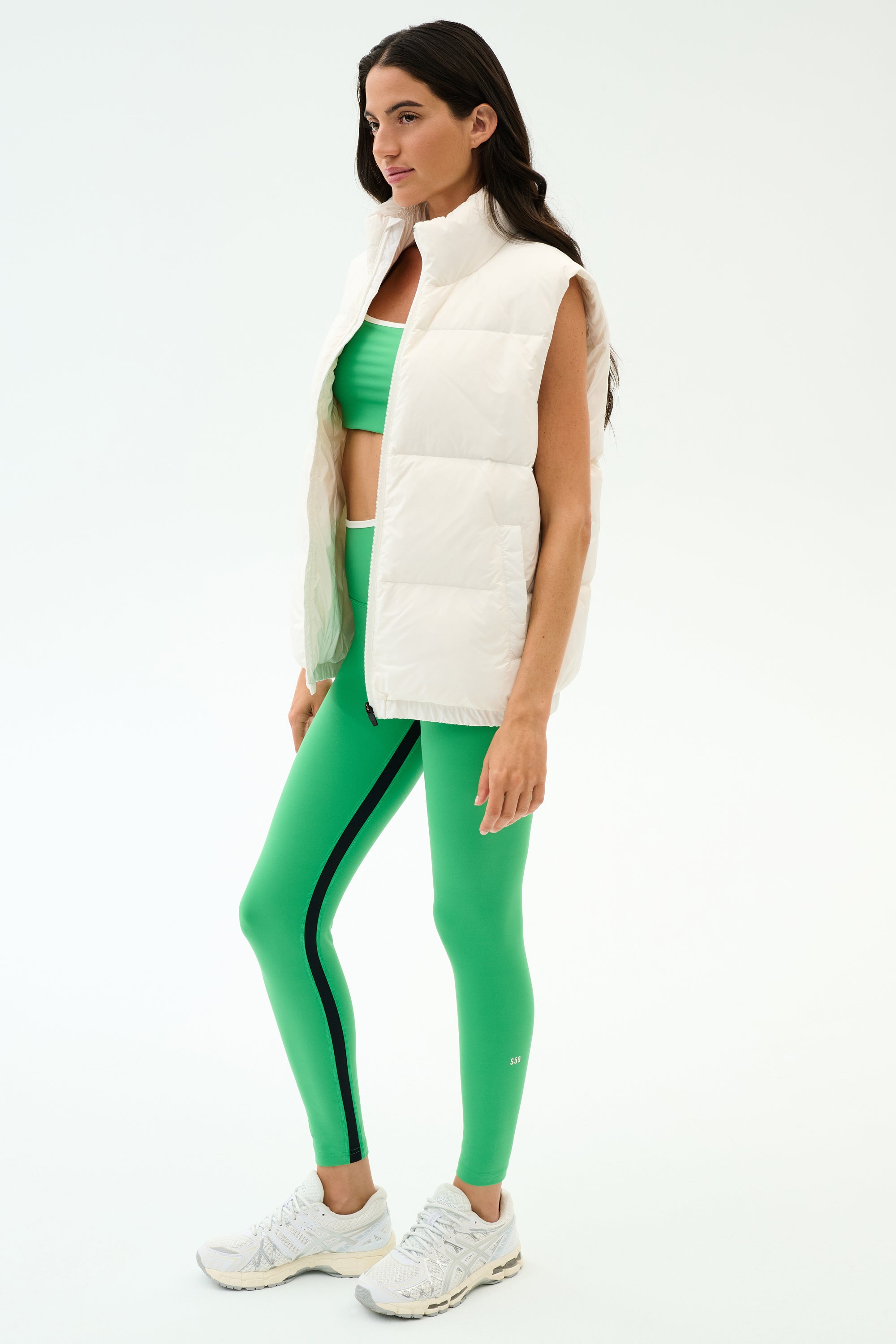 A woman stands in profile against a plain background wearing the SPLITS59 Summit Nylon Puffer Vest in white, paired with a green sports bra, green leggings with black stripes, and white athletic shoes—ideal for cool weather workouts.