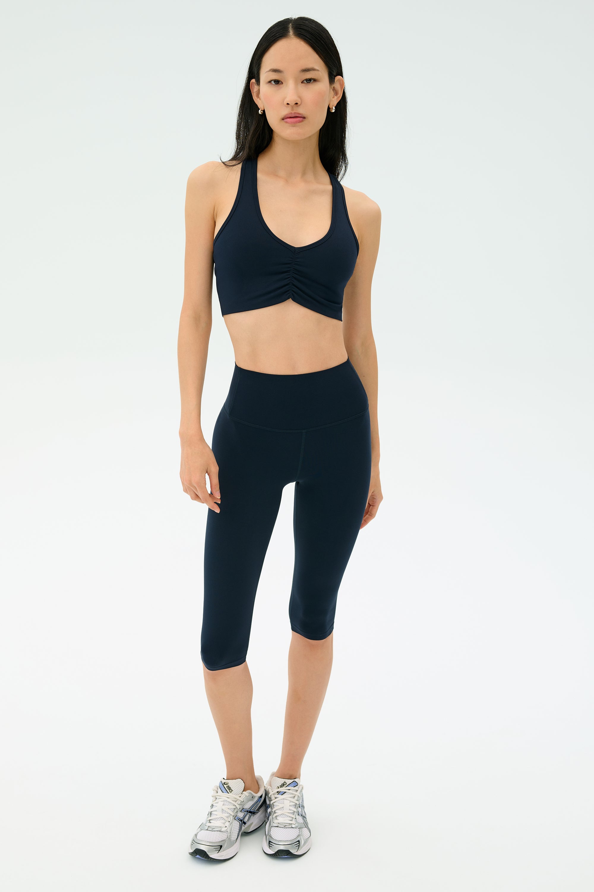 A woman faces forward wearing the SPLITS59 Airweight Bra with Ruching in Indigo, paired with matching navy capri leggings and silver athletic shoes against a plain light background.