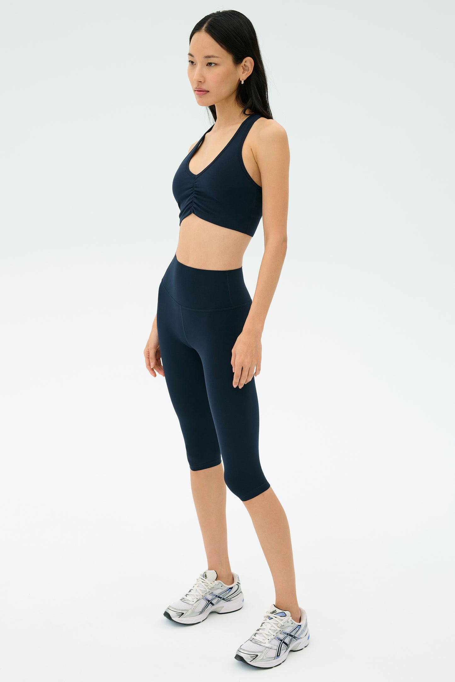 A woman poses against a plain background, wearing the SPLITS59 Airweight Bra with Ruching in Indigo and matching high-waisted capri leggings, paired with white running shoes.