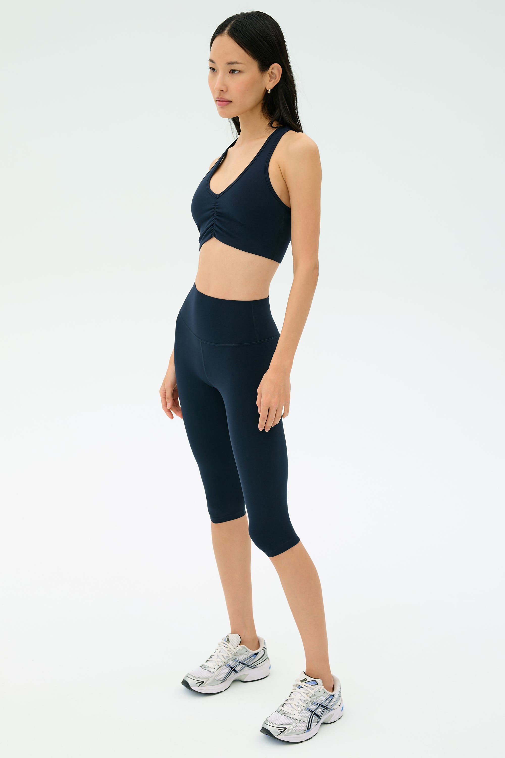 A woman poses against a plain background, wearing the SPLITS59 Airweight Bra with Ruching in Indigo and matching high-waisted capri leggings, paired with white running shoes.