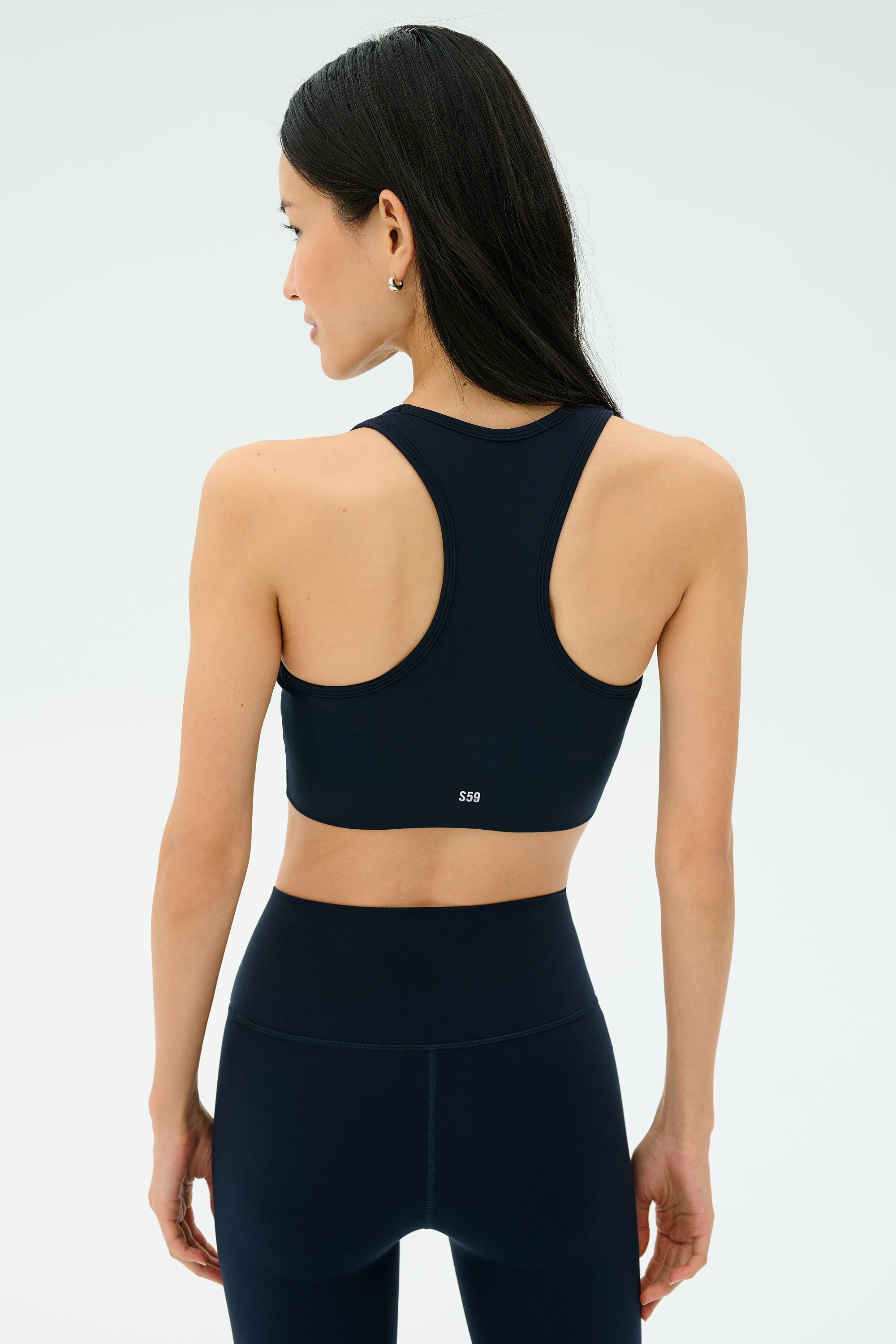Woman in black athletic wear stands facing away, showcasing the SPLITS59 Airweight Bra with Ruching in Indigo and high-waisted leggings against a plain light background.