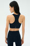Woman in black athletic wear stands facing away, showcasing the SPLITS59 Airweight Bra with Ruching in Indigo and high-waisted leggings against a plain light background.