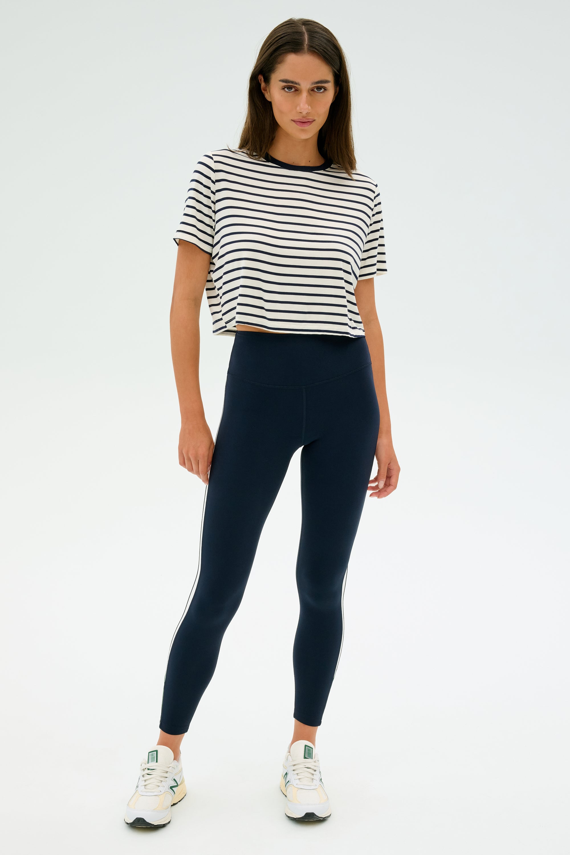 A woman stands against a plain background wearing the SPLITS59 Djuna Crop Ringer in Indigo Stripe, dark leggings, and sneakers, ready for her gym workouts.