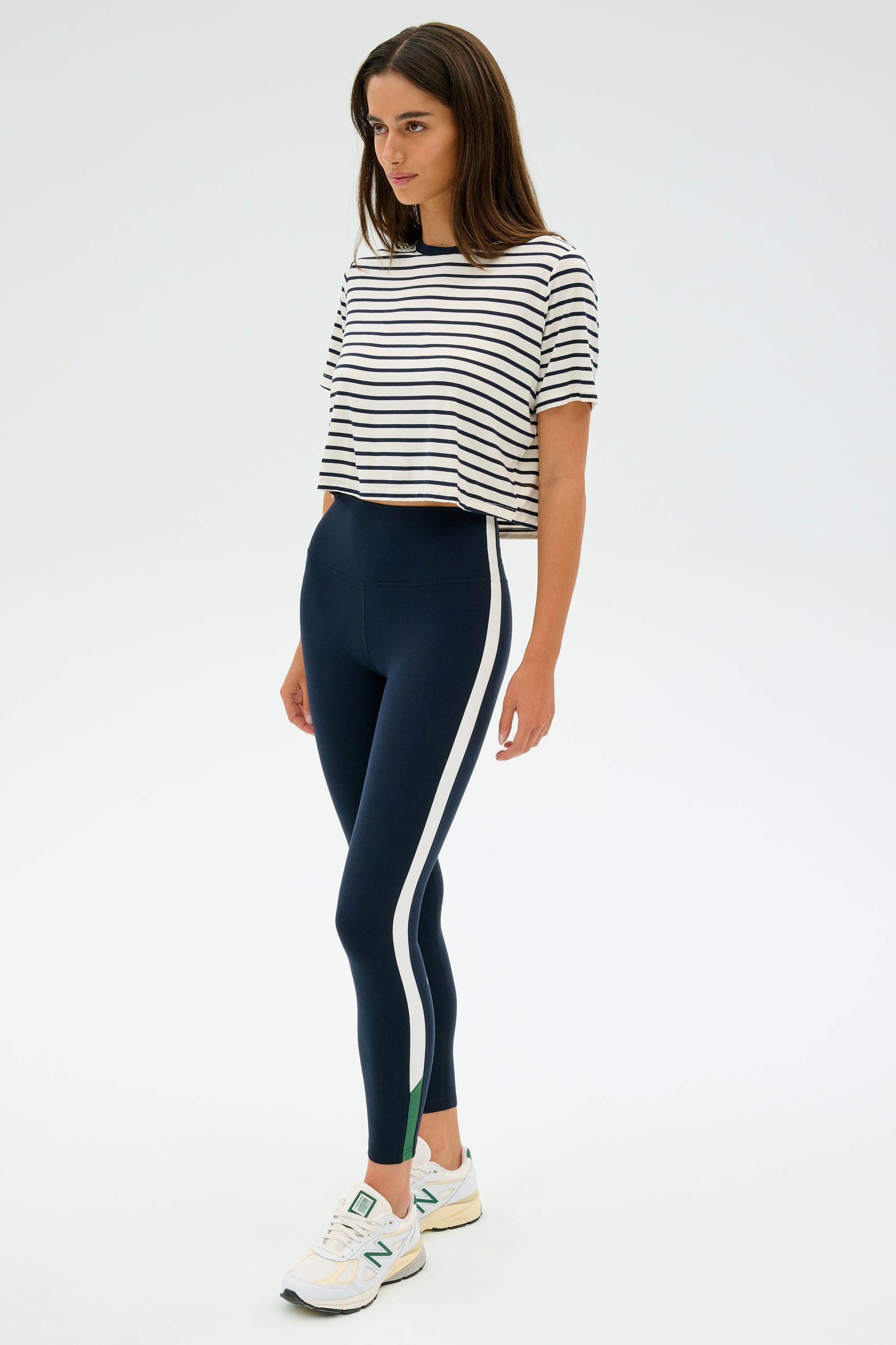 A woman stands wearing the Djuna Crop Ringer in Indigo Stripe by SPLITS59, navy leggings with white stripes, and white sneakers, ready for gym workouts against a plain background.