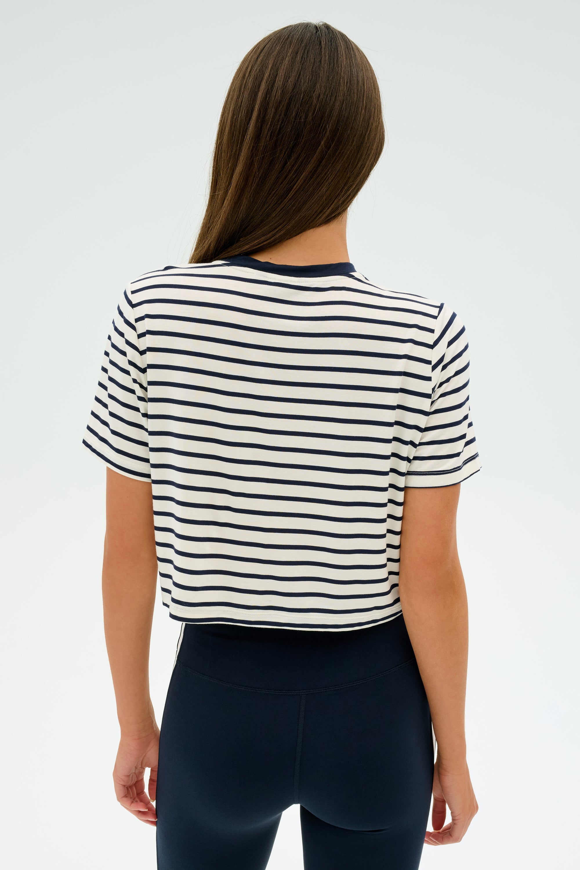 A long-haired fitness enthusiast wears SPLITS59's Djuna Crop Ringer in Indigo Stripe paired with black pants. With their back to the camera against a plain background, they exude a passion for gym workouts.