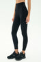 A person wears SPLITS59 Airweight Super High Waist 7/8 leggings in black with black athletic shoes, standing against a plain light background.