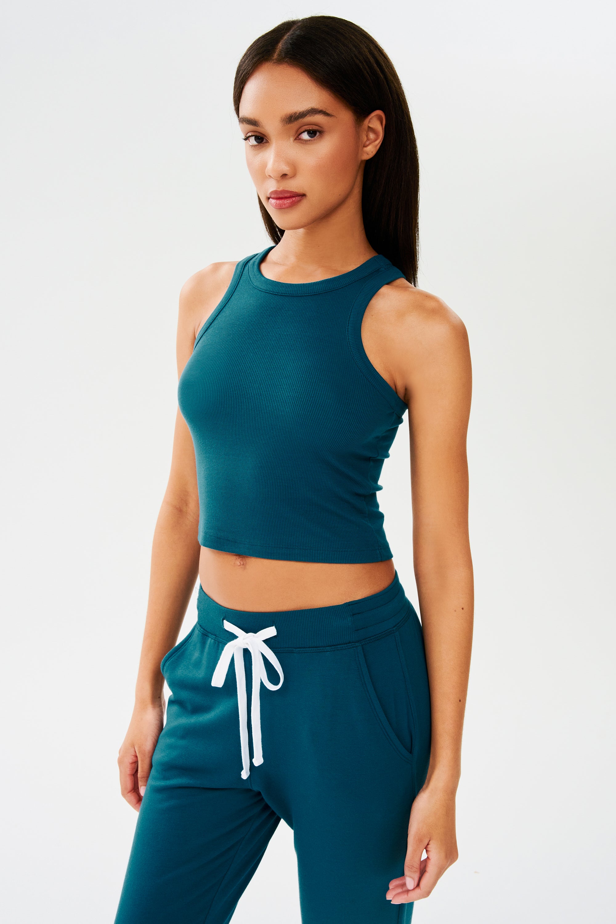 The model is wearing a SPLITS59 Kiki Rib Crop Tank in Peacock and joggers.