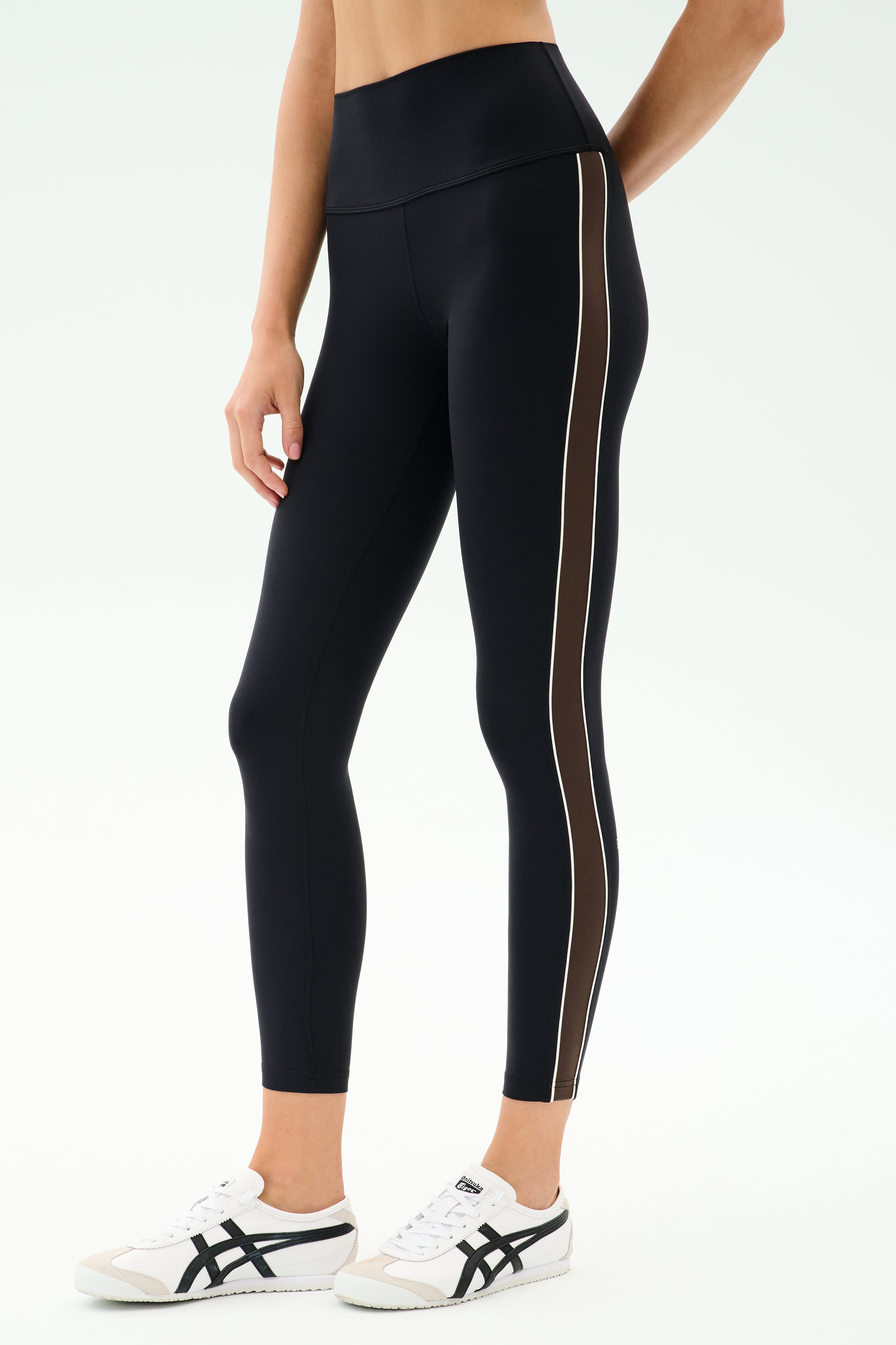 A person wears the SPLITS59 Margot Rigor Legging in Black/Dark Chocolate with brown and white side stripes, paired with white athletic shoes, standing against a plain background.