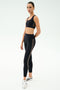 A person stands in a studio wearing the SPLITS59 Lexi Rigor Bra in Black/Dark Chocolate and black high-waisted leggings with white piping, paired with white sneakers, set against a plain light background—ideal for gym workouts.