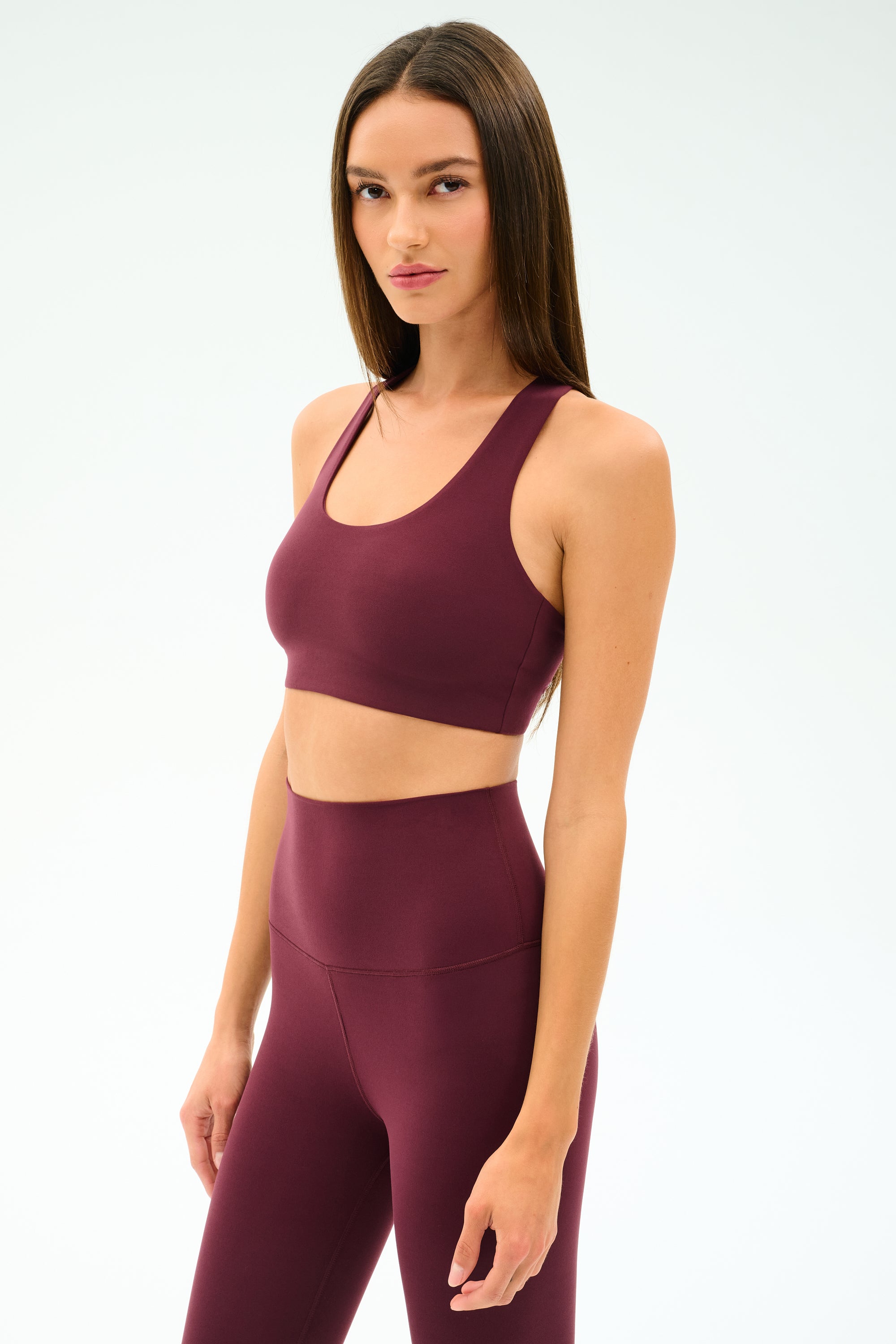 A woman wears the SPLITS59 Sara Airweight Bra in Dark Cherry with matching high-waisted leggings, standing against a plain light background.