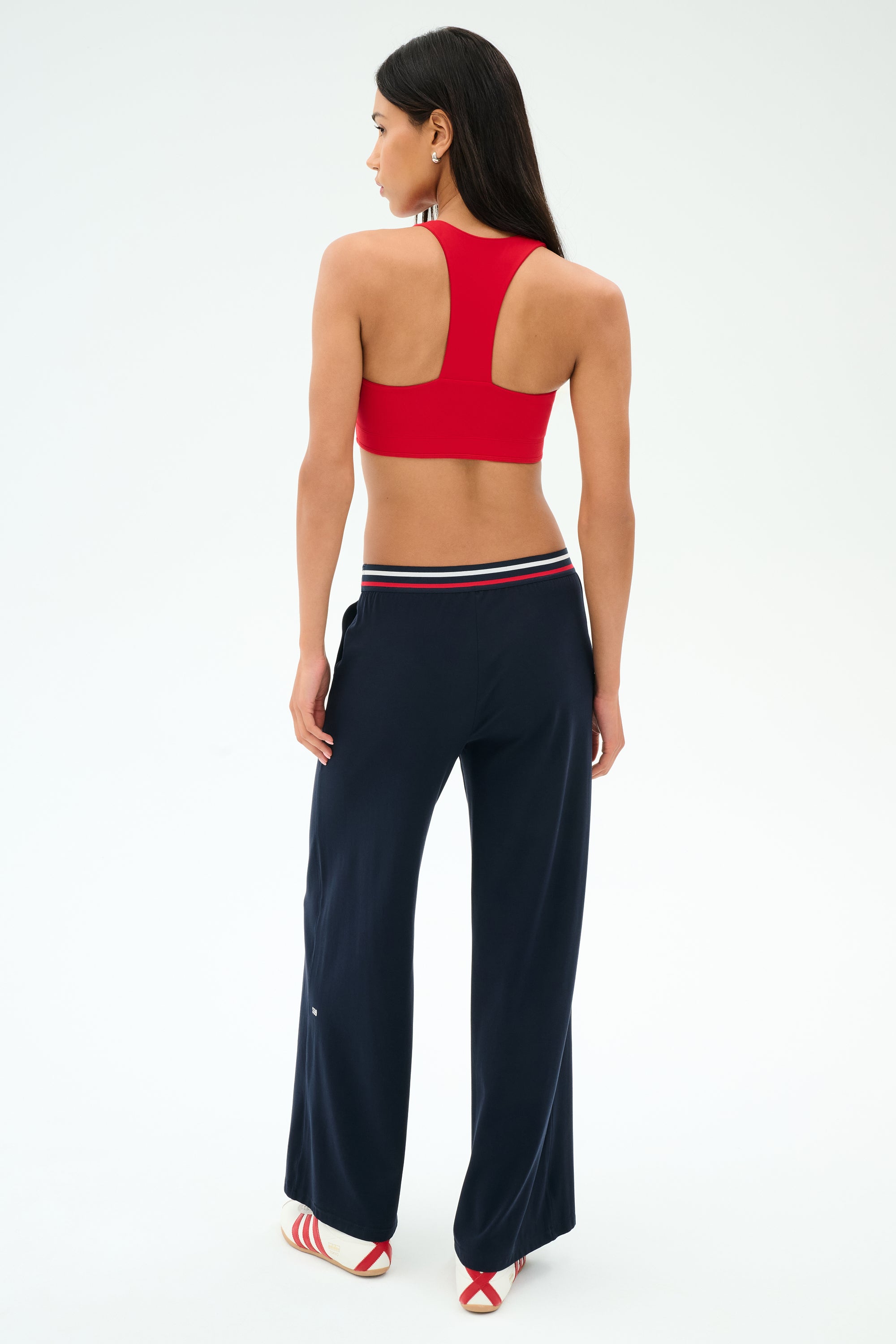 A woman faces away from the camera wearing a red racerback sports bra, SPLITS59 Michele Airweight Trouser in Indigo with a striped waistband, and red and white sandals against a plain backdrop.