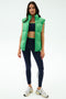 A woman wears the SPLITS59 Summit Nylon Puffer Vest in Grass over a navy sports bra, striped navy leggings, and white sneakers—ideal for cool weather workouts—shown against a plain light background.
