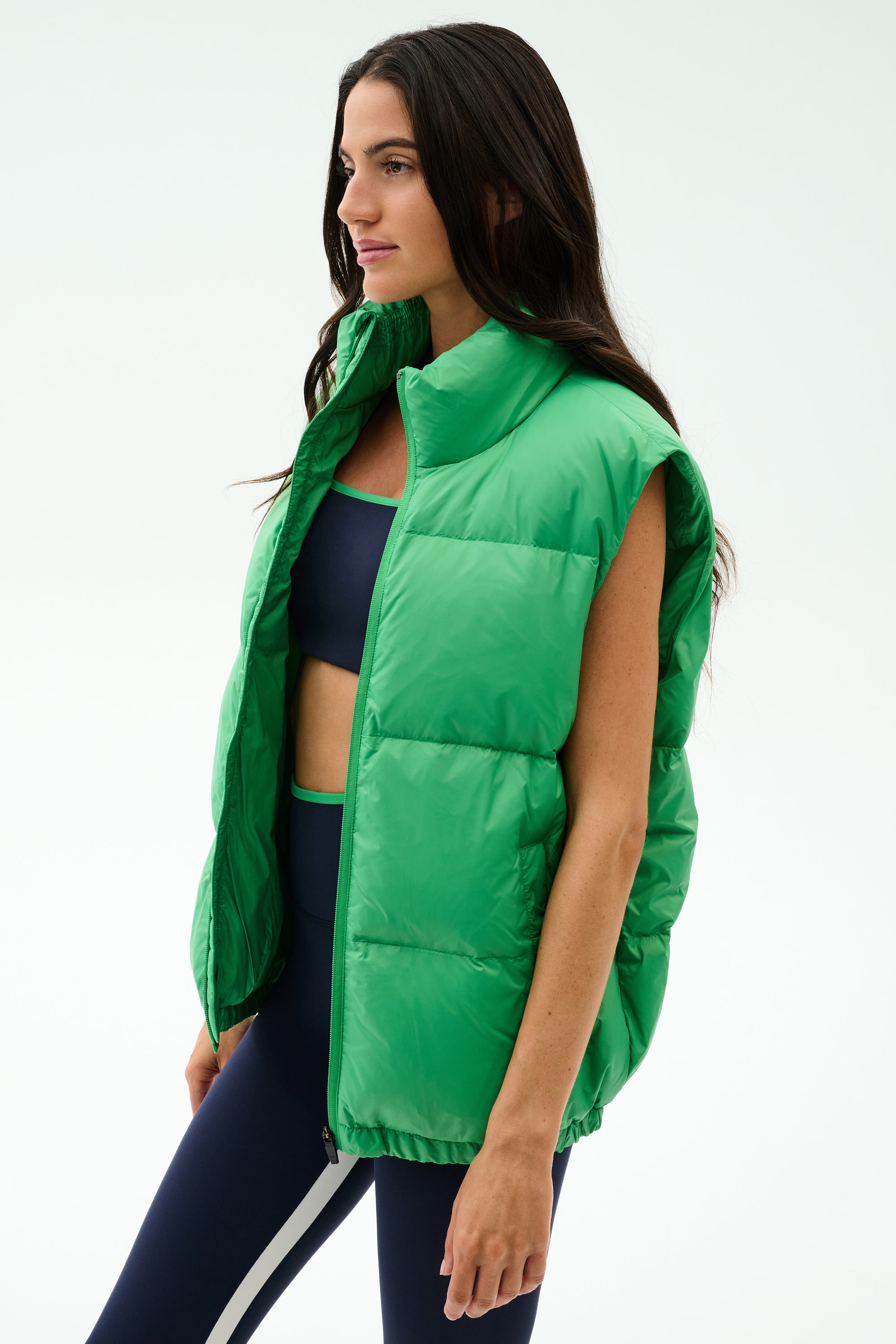 A woman wears the SPLITS59 Summit Nylon Puffer Vest in Grass over a navy sports bra and navy leggings with a white stripe, standing against a plain white background—ideal for cool weather workouts with its lightweight insulation.