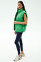 A woman wears the SPLITS59 Summit Nylon Puffer Vest in Grass for lightweight warmth, paired with navy leggings featuring a white stripe and white sneakers—ideal for cool weather workouts against a plain light backdrop.