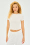 A curly-haired person stands against a white background in a short sleeve SPLITS59 Jennie Jersey Tee and white pants, ready for gym workouts.