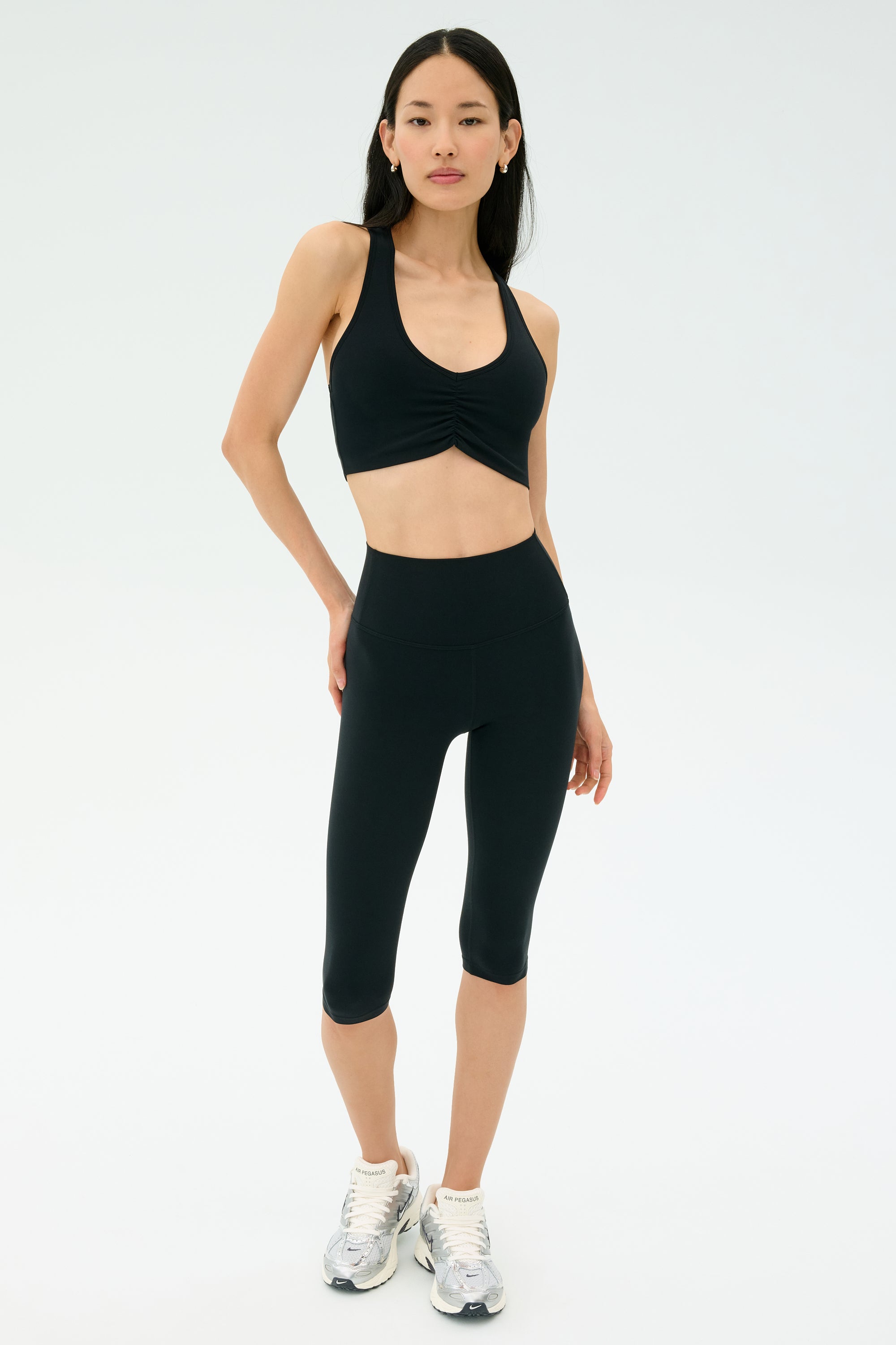 A person stands against a plain background wearing a black SPLITS59 Airweight Bra with Ruching, black high-waisted capri leggings, and gray running shoes.
