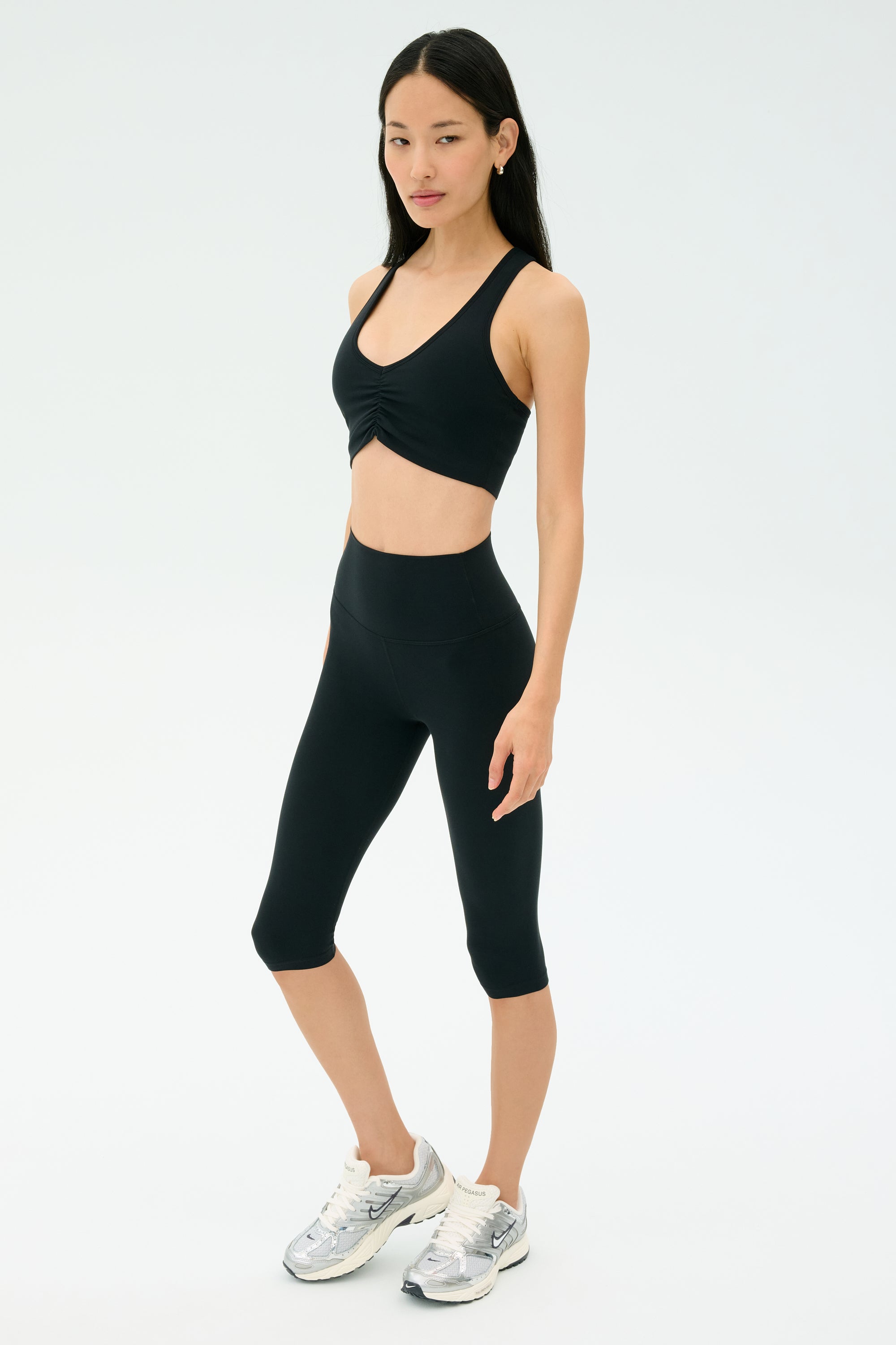 A woman stands against a plain background wearing the SPLITS59 Airweight Bra with Ruching in black, paired with black capri leggings and silver athletic shoes.