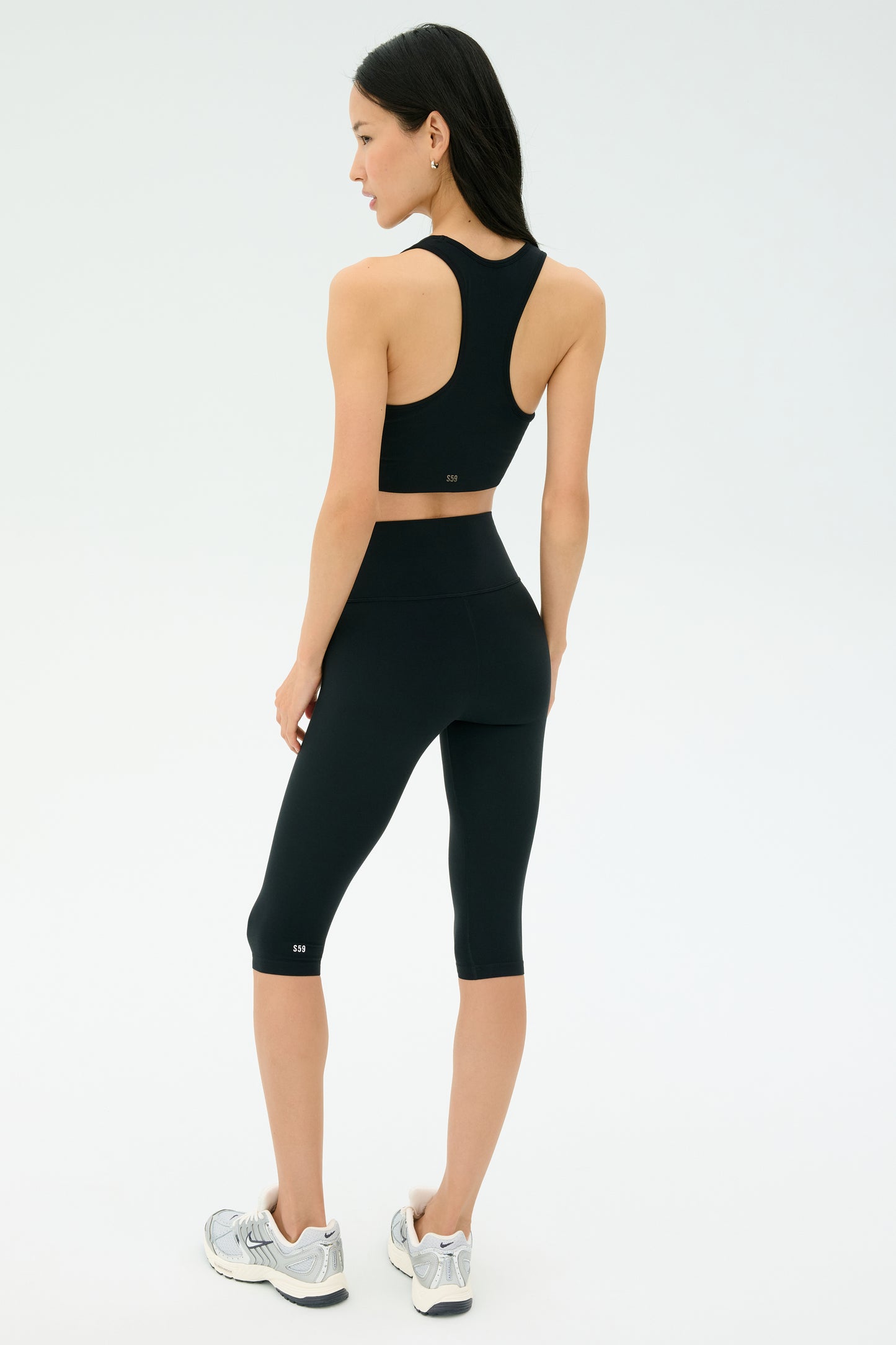 A woman stands facing away against a plain light background, wearing SPLITS59's Airweight High Waist 16" Legging in black, a matching black sports bra, and silver running shoes.