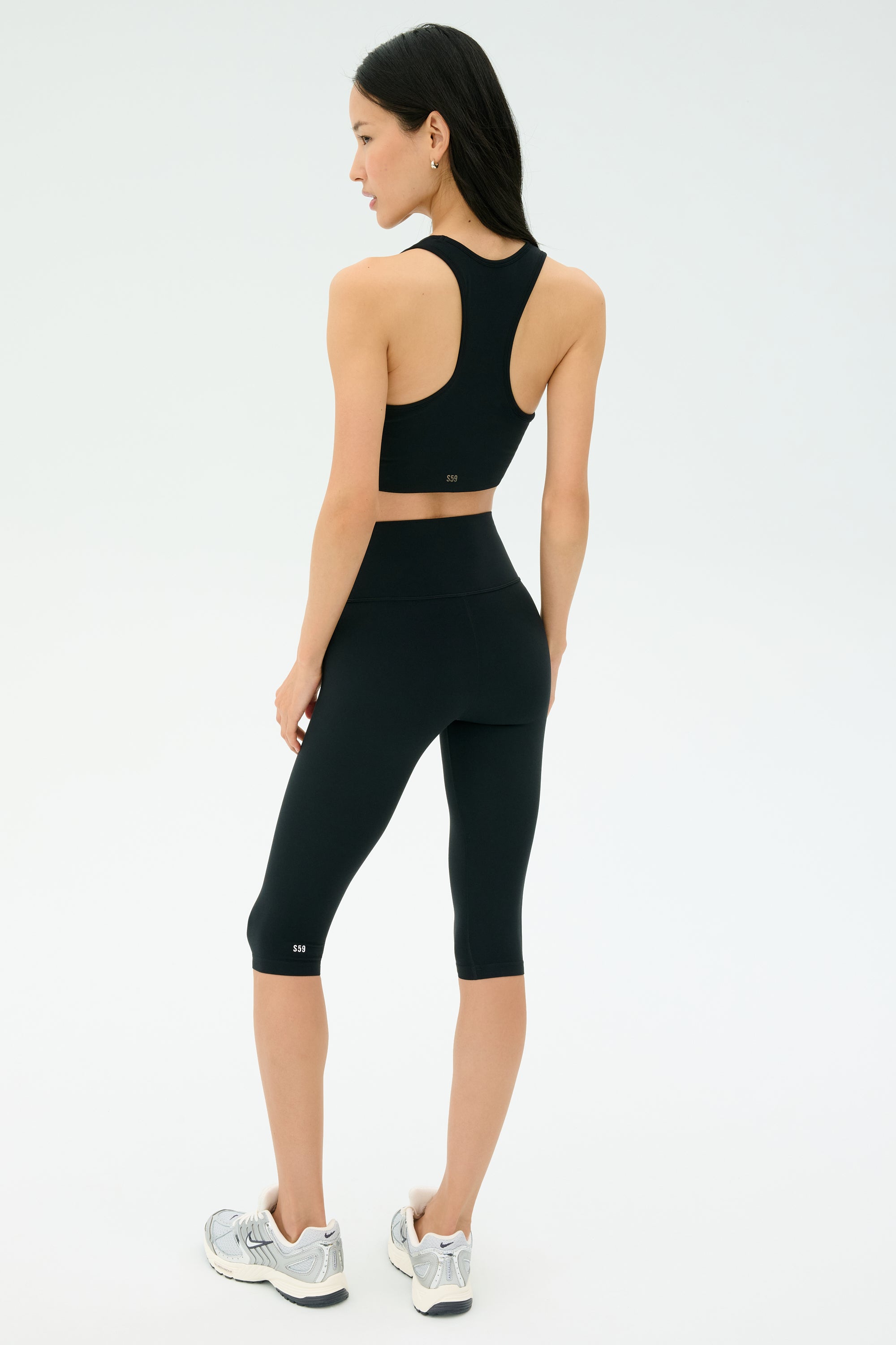 A woman stands facing away against a plain light background, wearing SPLITS59's Airweight High Waist 16" Legging in black, a matching black sports bra, and silver running shoes.