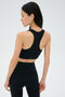 A woman in black athletic wear stands facing away, highlighting the back of the SPLITS59 Airweight Bra with Ruching and matching leggings, both crafted from ultra-light Airweight performance fabric, against a plain light background.