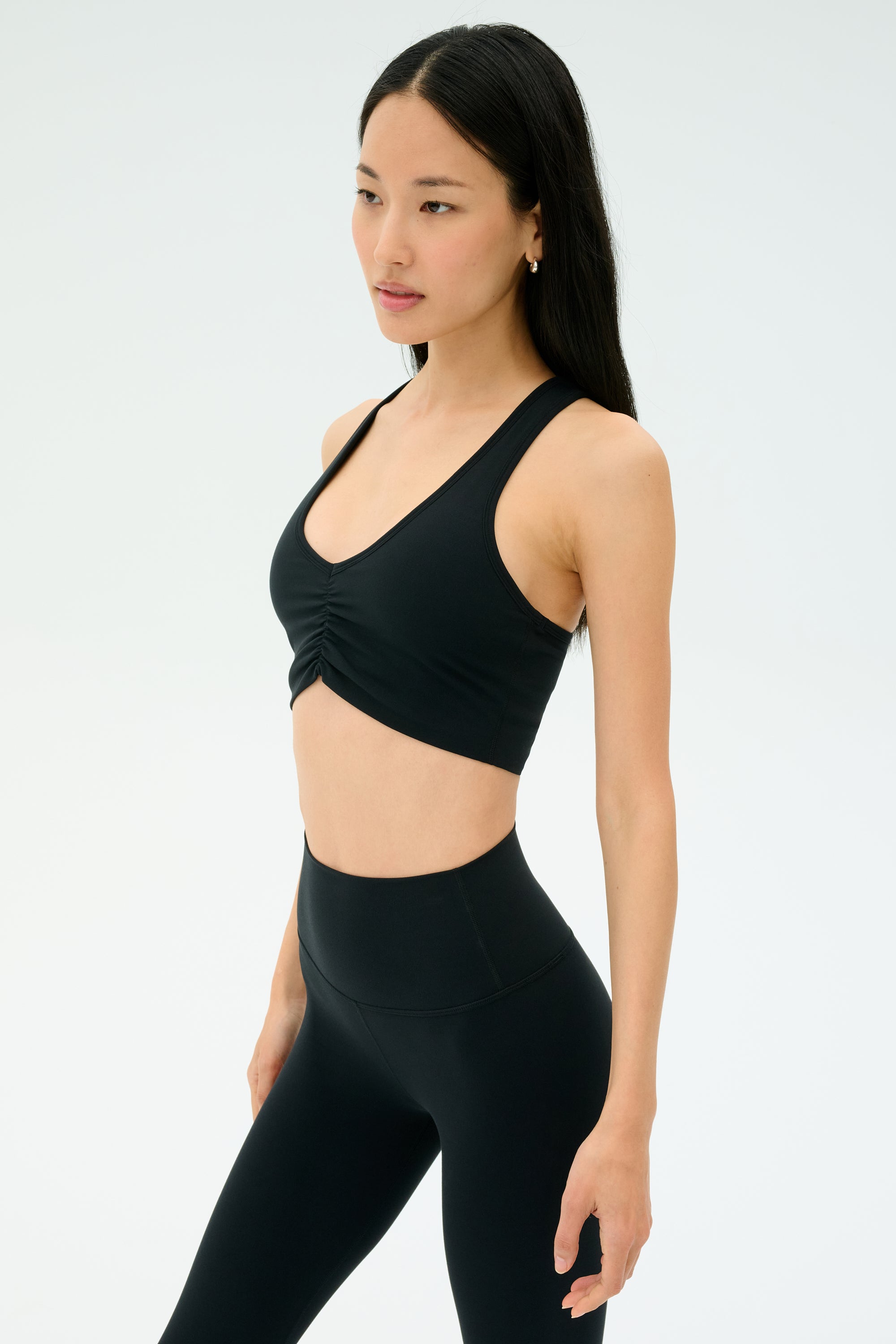 A woman wears the SPLITS59 Airweight Bra with Ruching in black and matching high-waisted black leggings, both in ultra-light Airweight performance fabric, standing against a plain light background.