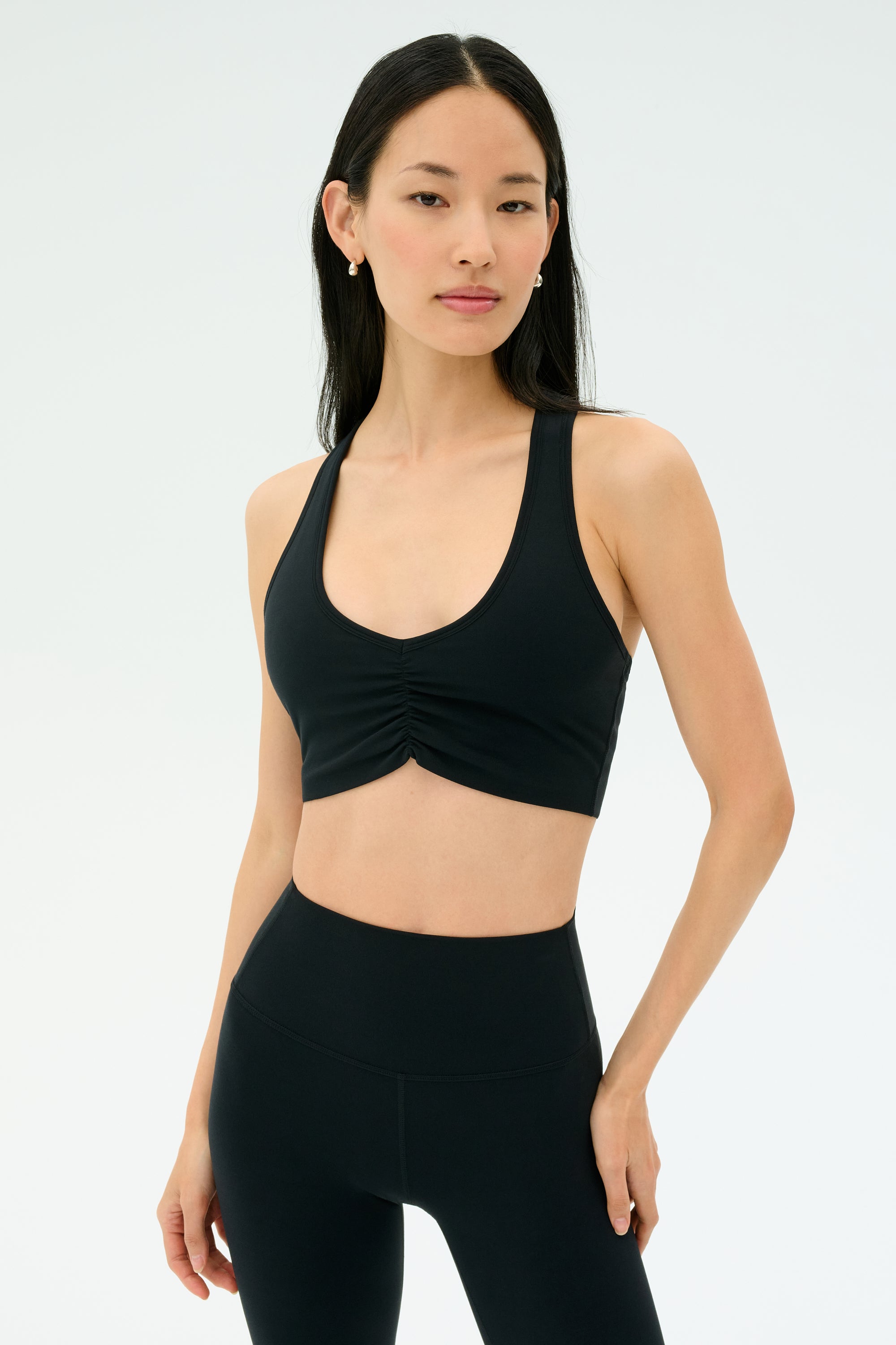 A woman stands against a plain background, wearing the SPLITS59 Airweight Bra with Ruching in black and high-waisted leggings, looking at the camera with a neutral expression.