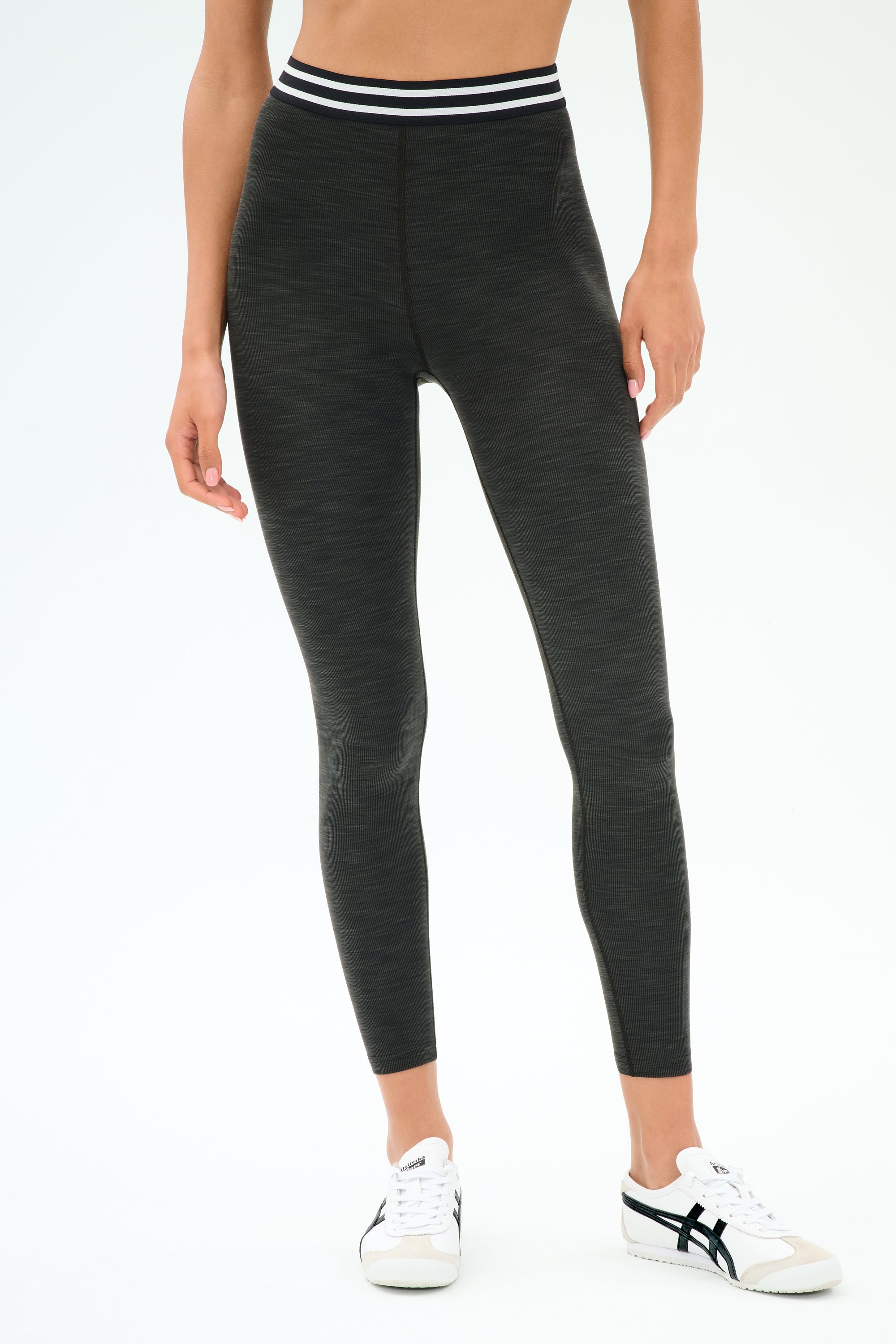 A person wears SPLITS59's Bailey Active Rib High Waist 7/8 leggings in Heather Graphite, featuring a striped waistband, paired with white sneakers—ideal for gym workouts—set against a plain white background.