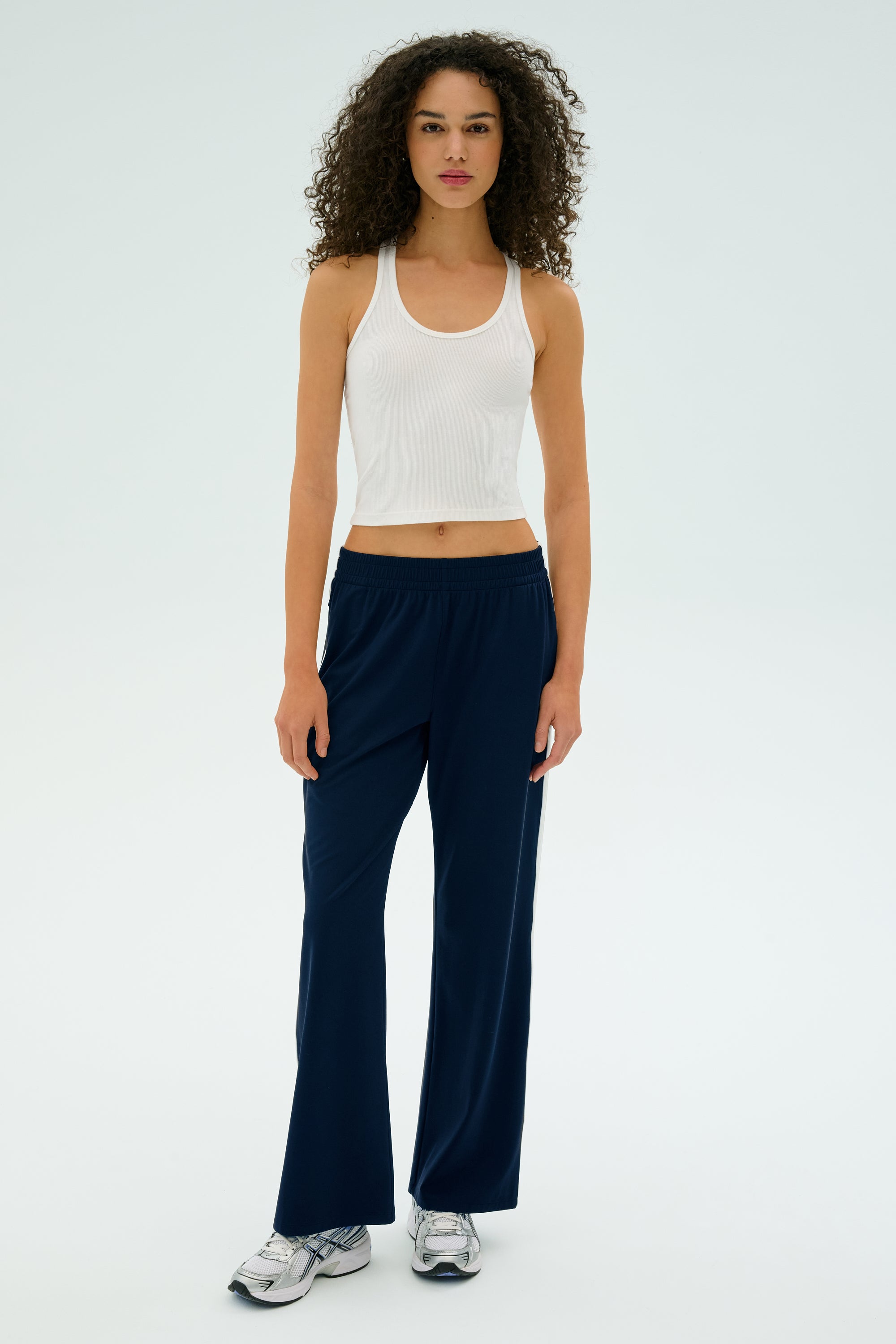 A person with curly hair stands against a light background, wearing a white tank top, SPLITS59's Maxie Rigor Track Pant in Indigo/White, and sneakers.