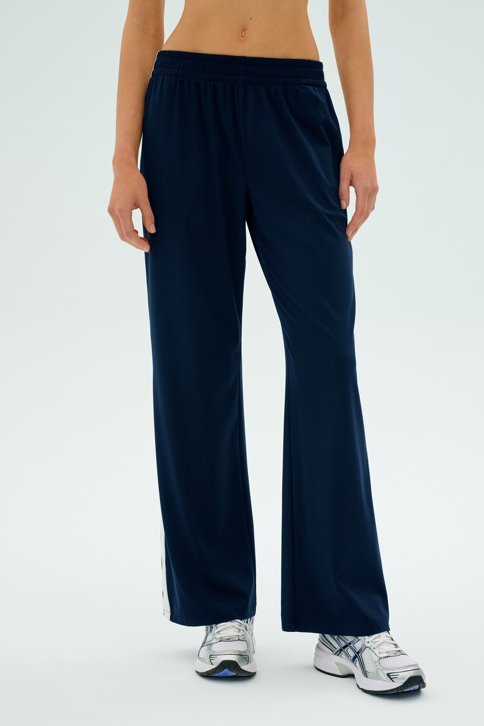 A person wearing SPLITS59's Maxie Rigor Track Pant in Indigo/White with a wide-leg silhouette, paired with silver sneakers, stands against a light background.