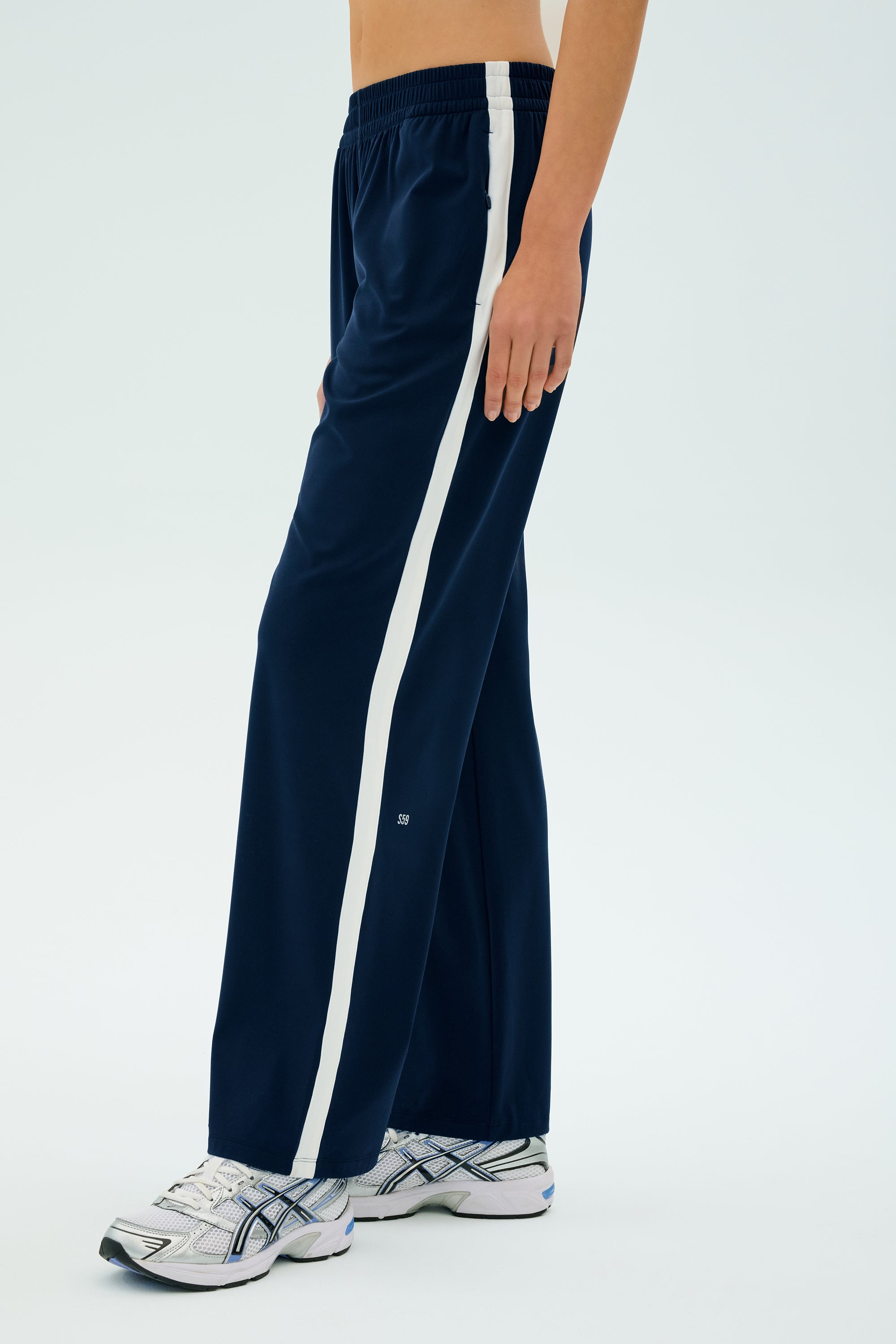 Someone in SPLITS59's Maxie Rigor Track Pant in Indigo/White, featuring white stripes and a wide-leg silhouette, paired with white sneakers, stands against a plain background.