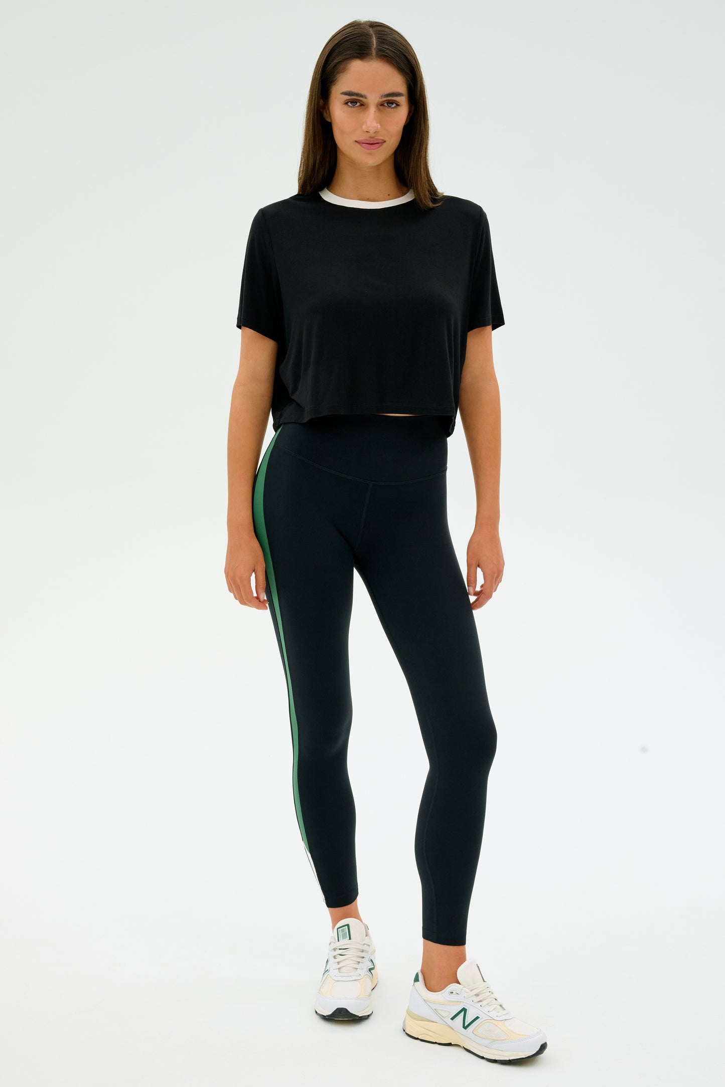 A woman in the SPLITS59 Djuna Crop Ringer - Black/White and green-striped leggings, wearing white sneakers, stands against a white backdrop, capturing the essence of CrossFit and gym workouts.