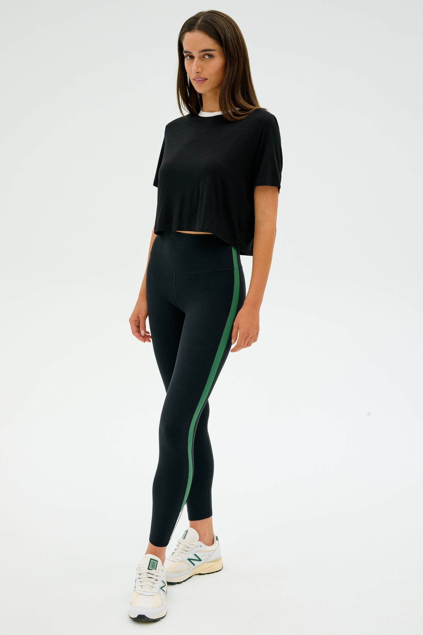 A person wearing the SPLITS59 Djuna Crop Ringer in Black/White, paired with black leggings featuring a green stripe and white sneakers, stands against a plain background, capturing the essence of CrossFit workouts.