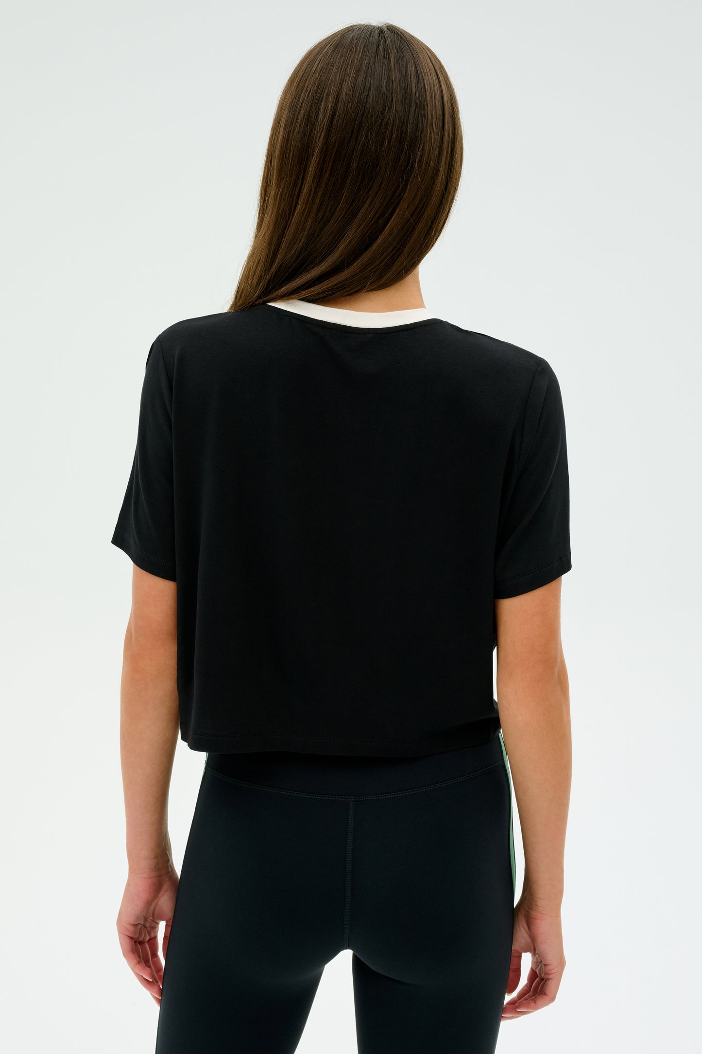 Facing away with long hair, the person is poised for an intense CrossFit session against a plain backdrop, wearing SPLITS59's Djuna Crop Ringer - Black/White T-shirt and black leggings.