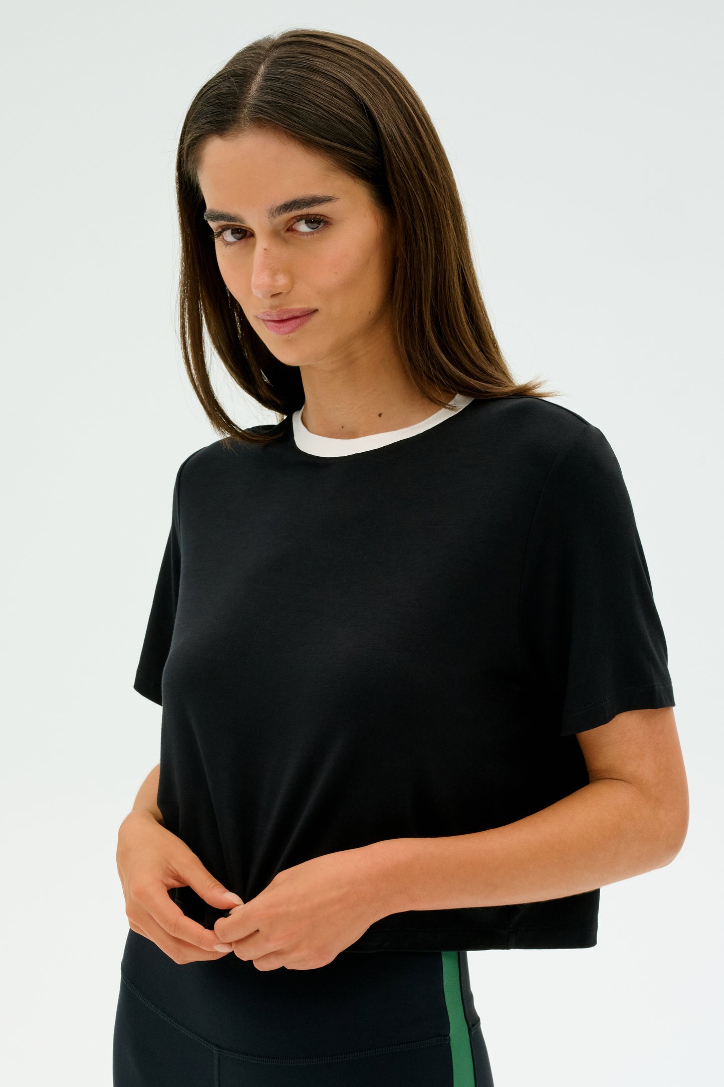 Sporting the SPLITS59 Djuna Crop Ringer in black/white, a person with long brown hair stands against a plain background. Their neutral gaze and determined stance reflect the resilience ingrained in CrossFit enthusiasts.