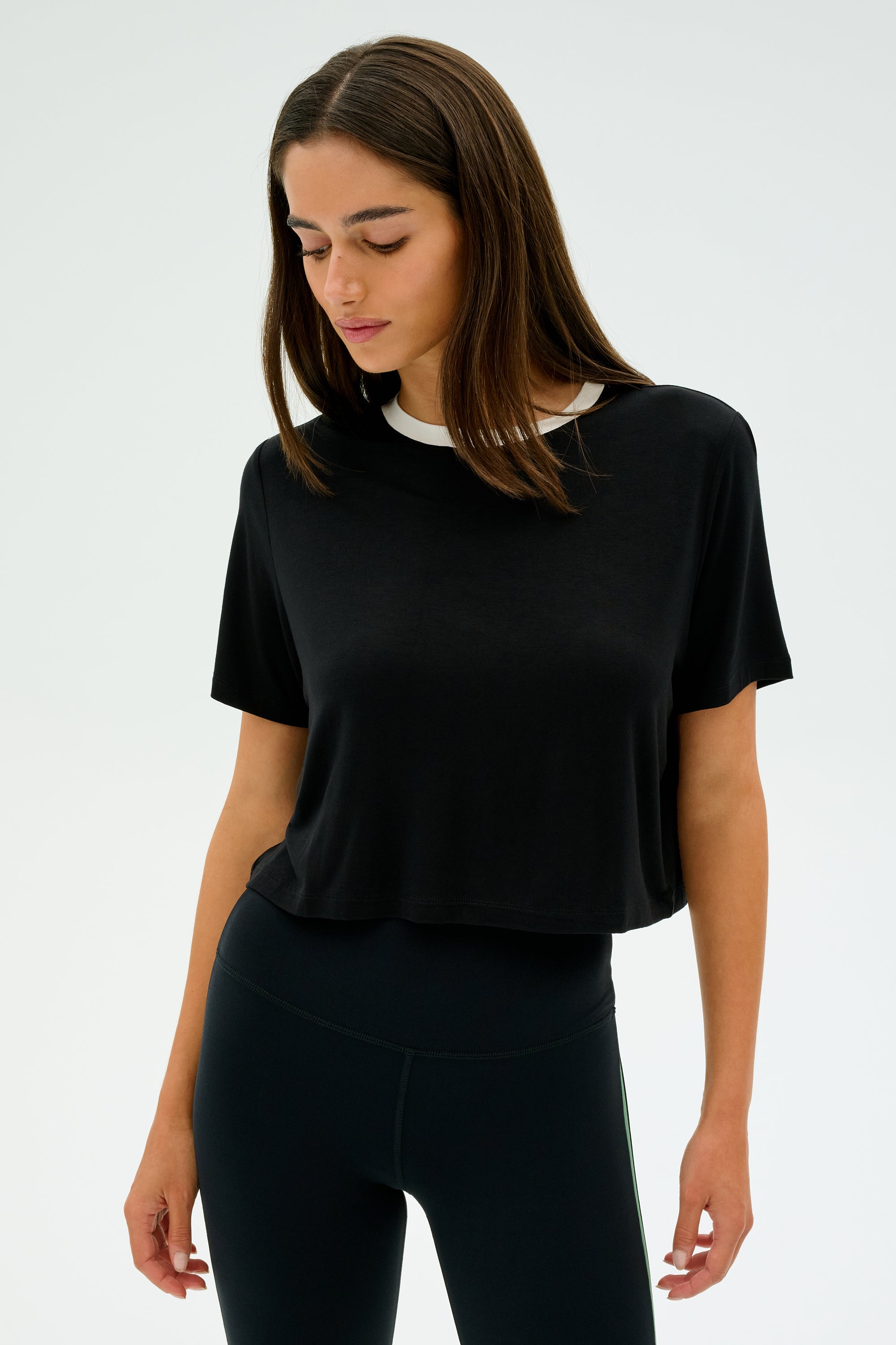 A person with long hair, poised against a plain white background, embodies the essence of gym workouts in the Djuna Crop Ringer in Black/White by SPLITS59 teamed with leggings.