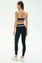 A woman stands facing away, ready to work out in SPLITS59's Mason High Waist Rigor Full Length leggings in Black/White and a black sports bra with white straps, paired with white sneakers against a plain background.