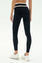 Model wearing SPLITS59 Mason High Waist Rigor Full Length black leggings with a white waistband and white sneakers, standing against a plain light background, facing away from the camera.