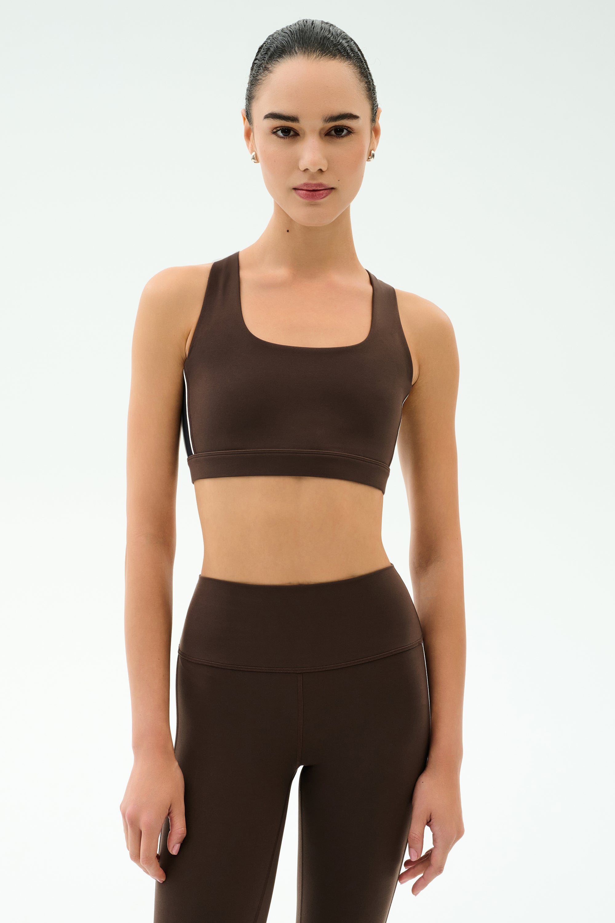 A woman wears the SPLITS59 Lexi Rigor Bra in Dark Chocolate/Black, paired with high-waisted leggings, standing against a plain light background and ready for high impact workouts.