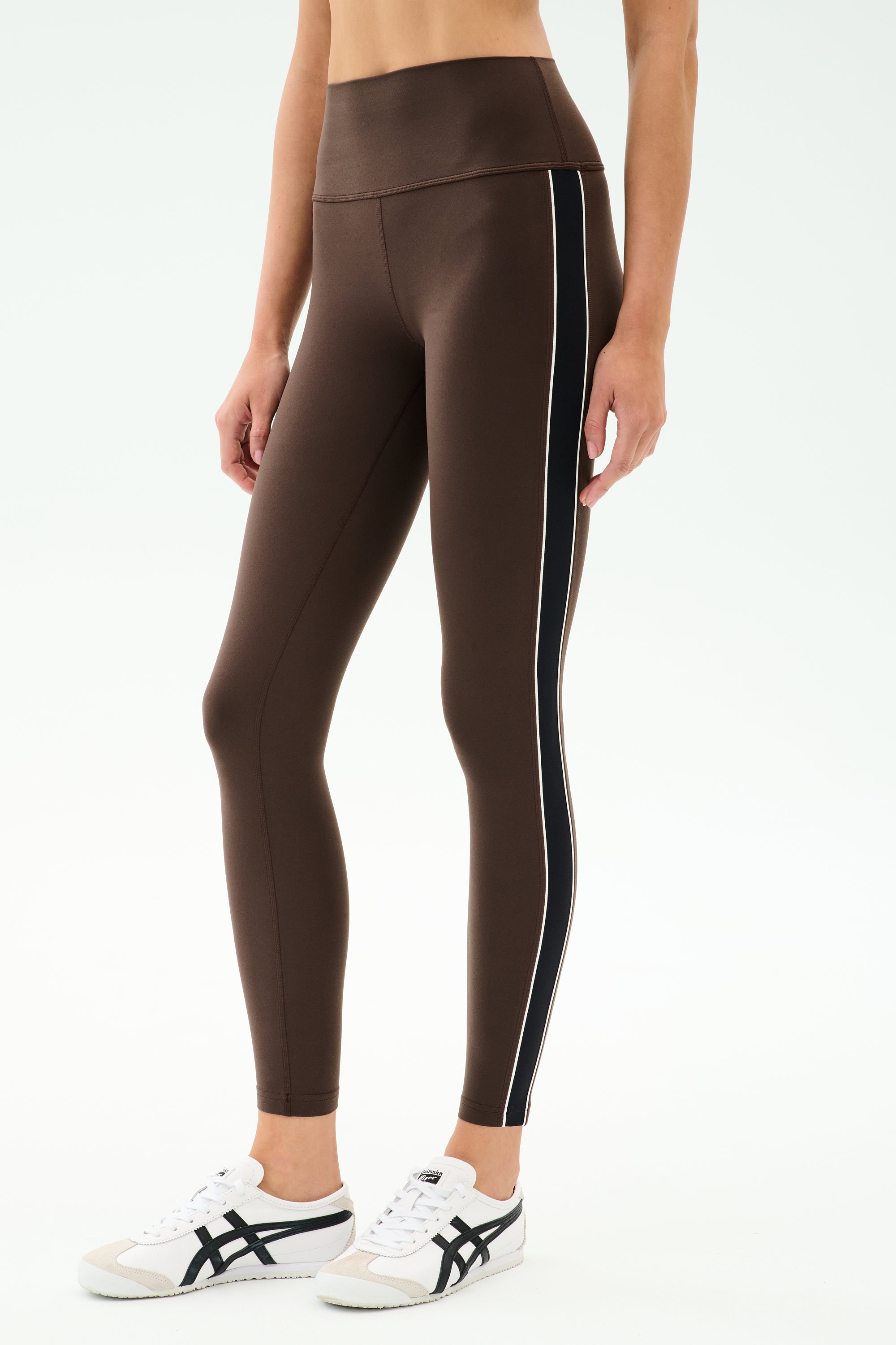 A person wearing SPLITS59 Margot Rigor Legging in Dark Chocolate/Black with a white side stripe and white sneakers with black accents stands against a plain background.