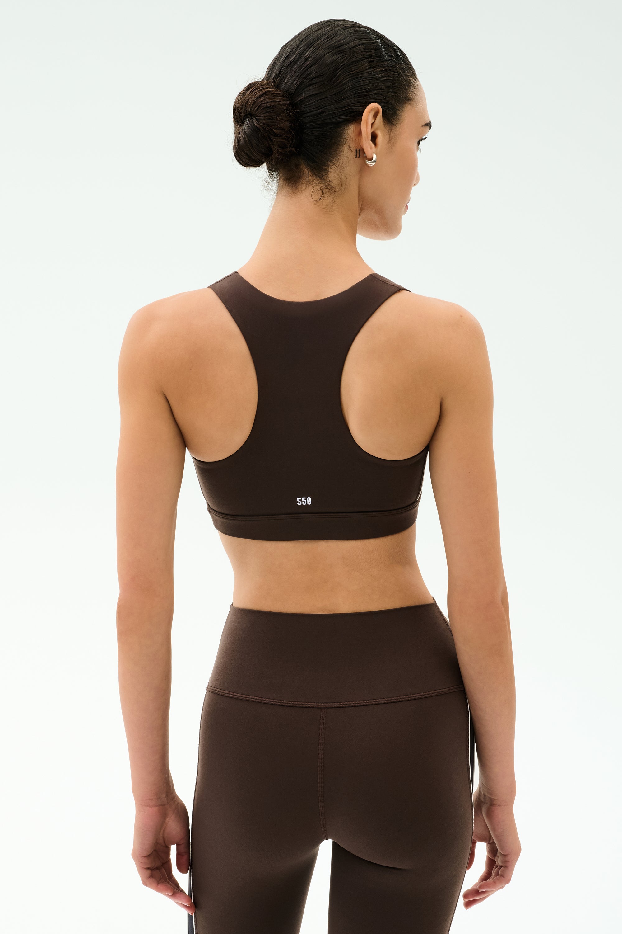 A woman with dark hair in a bun wears the SPLITS59 Lexi Rigor Bra in Dark Chocolate/Black, ideal for gym workouts, paired with matching high-waisted leggings and shown from the back against a plain light background.