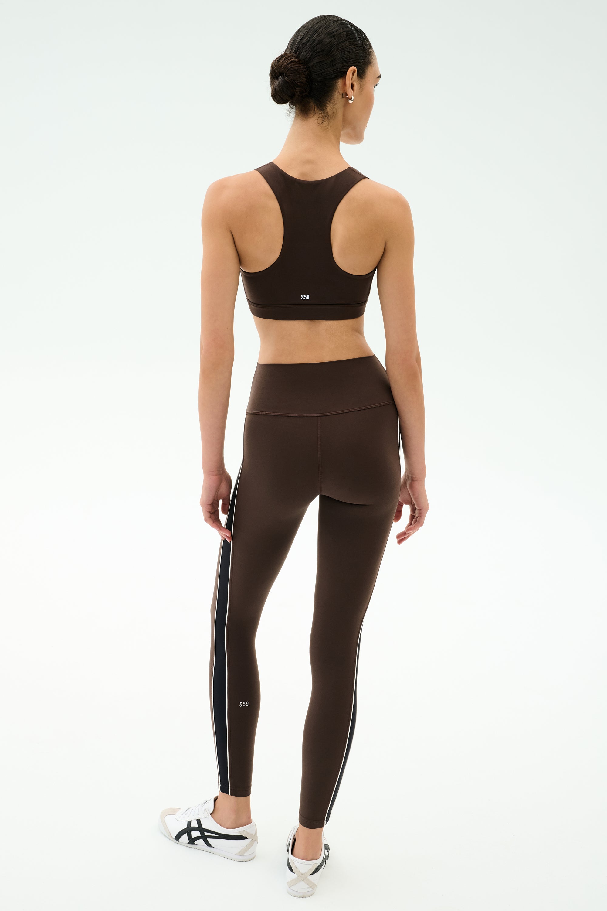 A woman faces away, wearing the SPLITS59 Lexi Rigor Bra in Dark Chocolate/Black and matching side-striped leggings, plus white sneakers with black accents, ready for high-impact workouts against a plain light background.