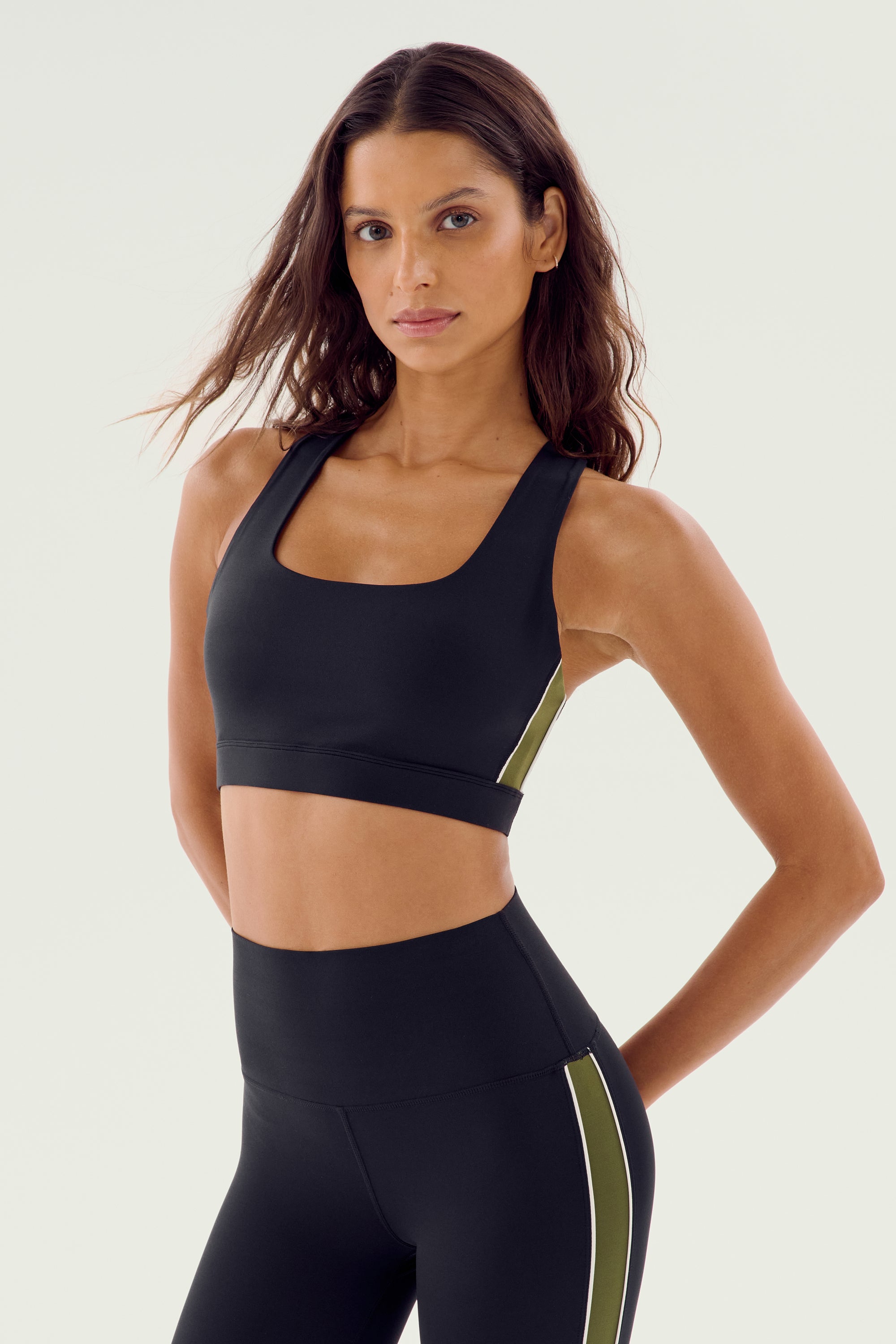 A person stands facing the camera, wearing a black Lexi Rigor Bra - Black/Olive by SPLITS59 and black nylon leggings with green and white stripes on the sides.