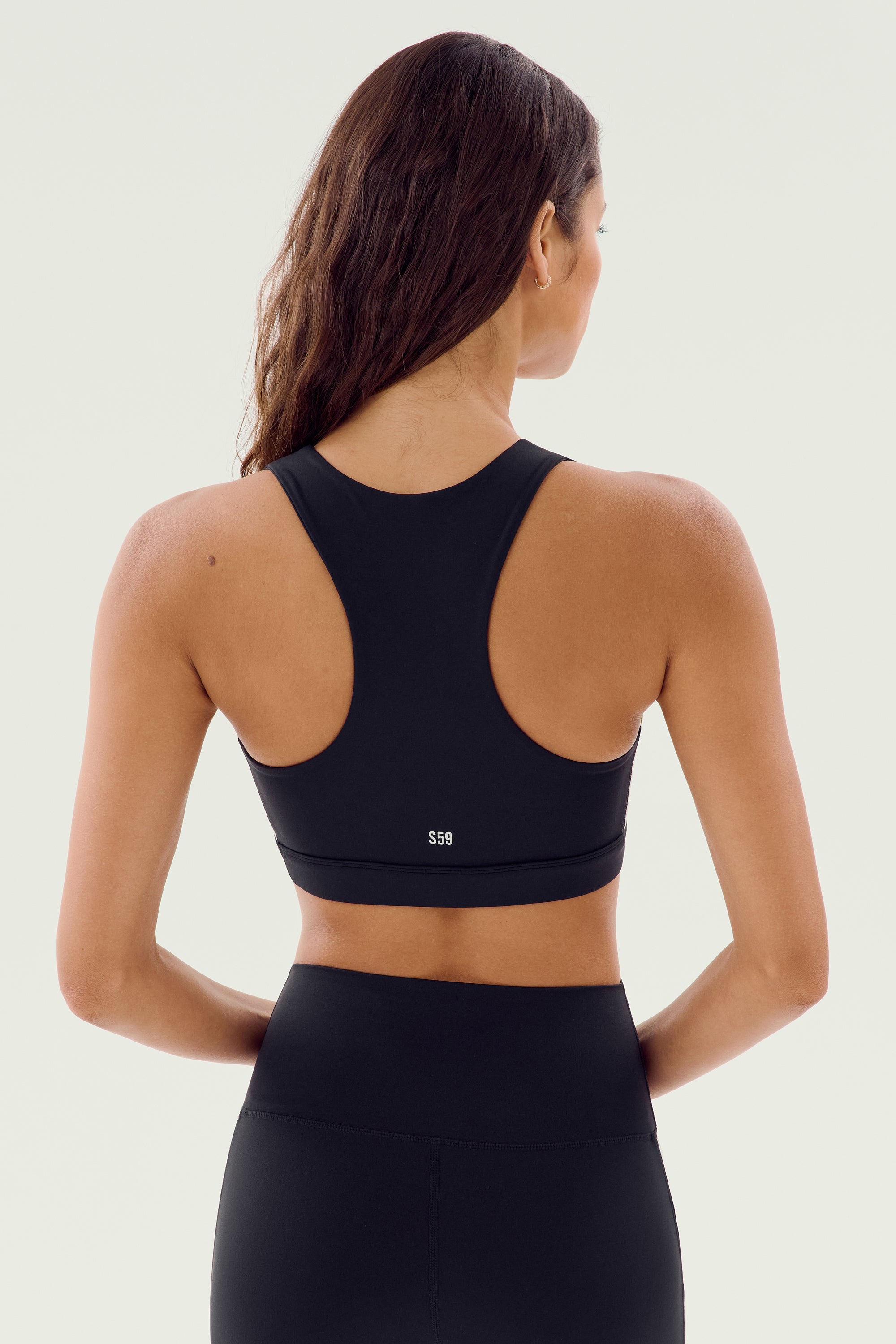 A woman with long brown hair is wearing a Lexi Rigor Bra in Black/Olive by SPLITS59 and matching spandex leggings, facing away from the camera against a plain background.