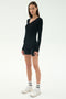 A woman stands on a white background wearing the SPLITS59 Louise Rib Henley in black, paired with black shorts, white athletic shoes, and white socks.