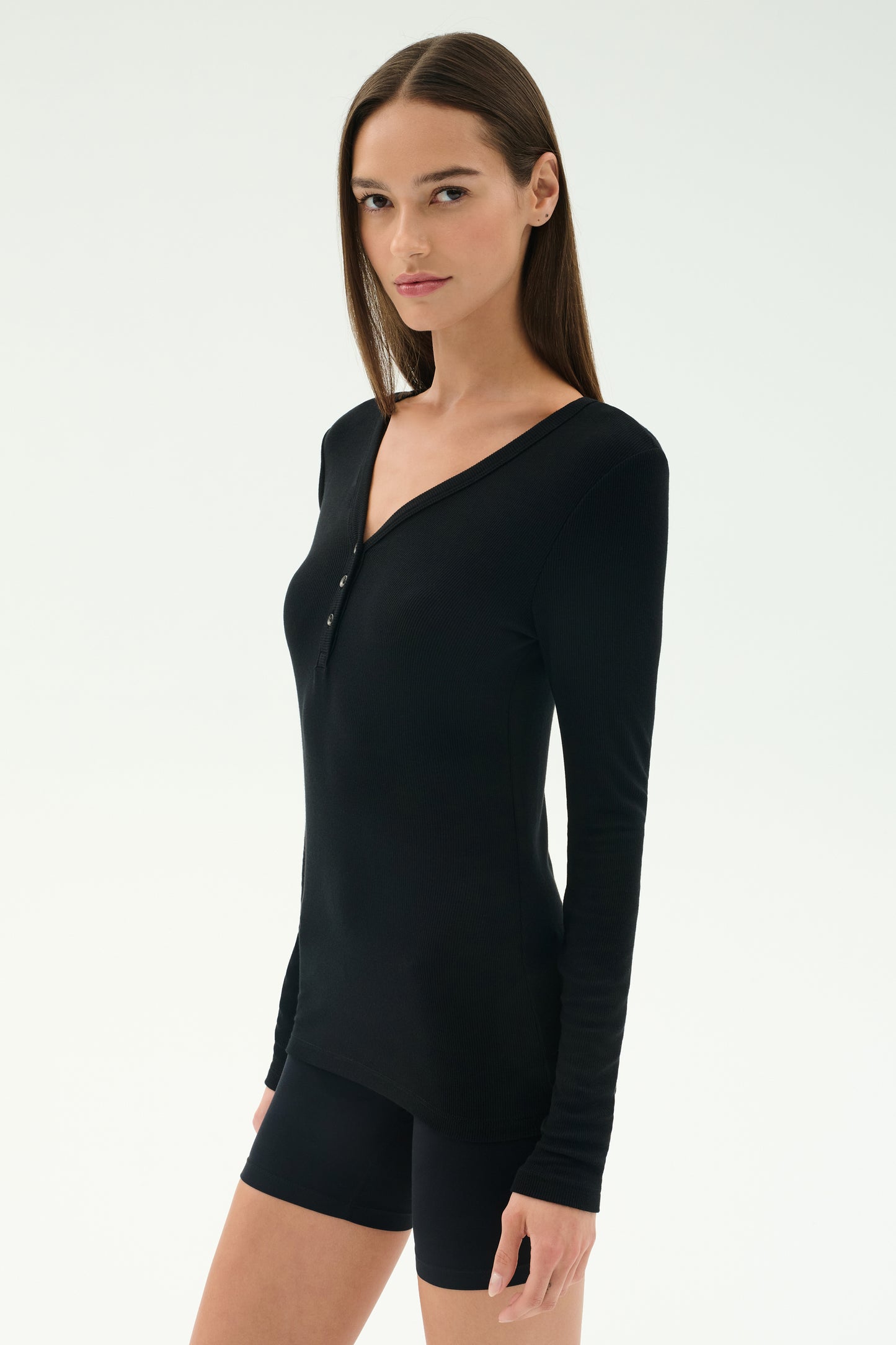 A woman with straight brown hair wears the SPLITS59 Louise Rib Henley in black and black shorts, standing against a plain white background.