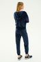 The individual with long blond hair is standing with arms crossed, back to the camera, wearing a navy blue hoodie and matching SPLITS59 Rhys Velour Jogger in Indigo, paired with black New Balance sneakers.